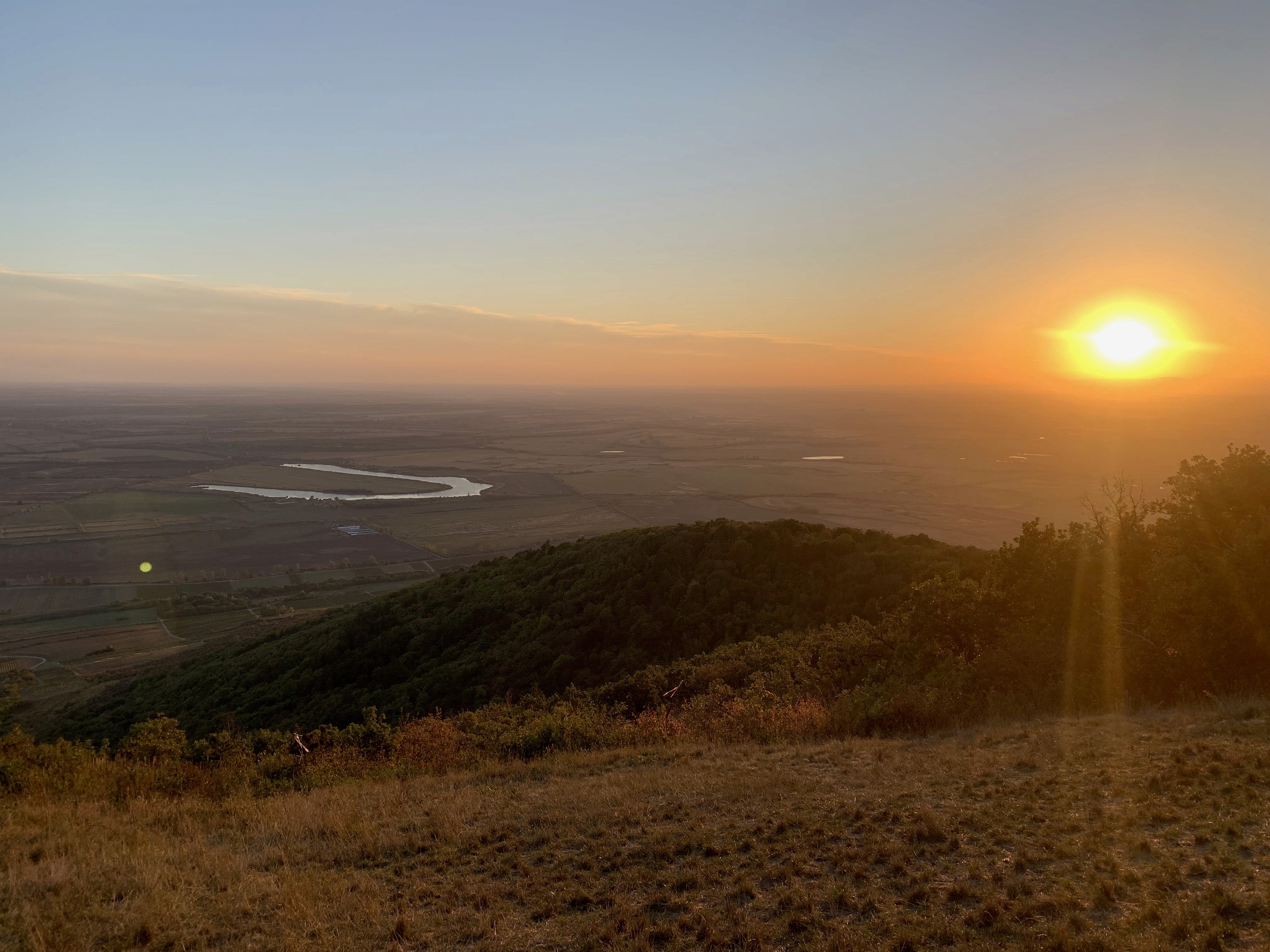 Lélegzetelállító naplemente aranyfényben fürdet egy nyugodt tájat, ahol kanyargó folyó és zöld dombok rajzolódnak ki a tiszta ég alatt.