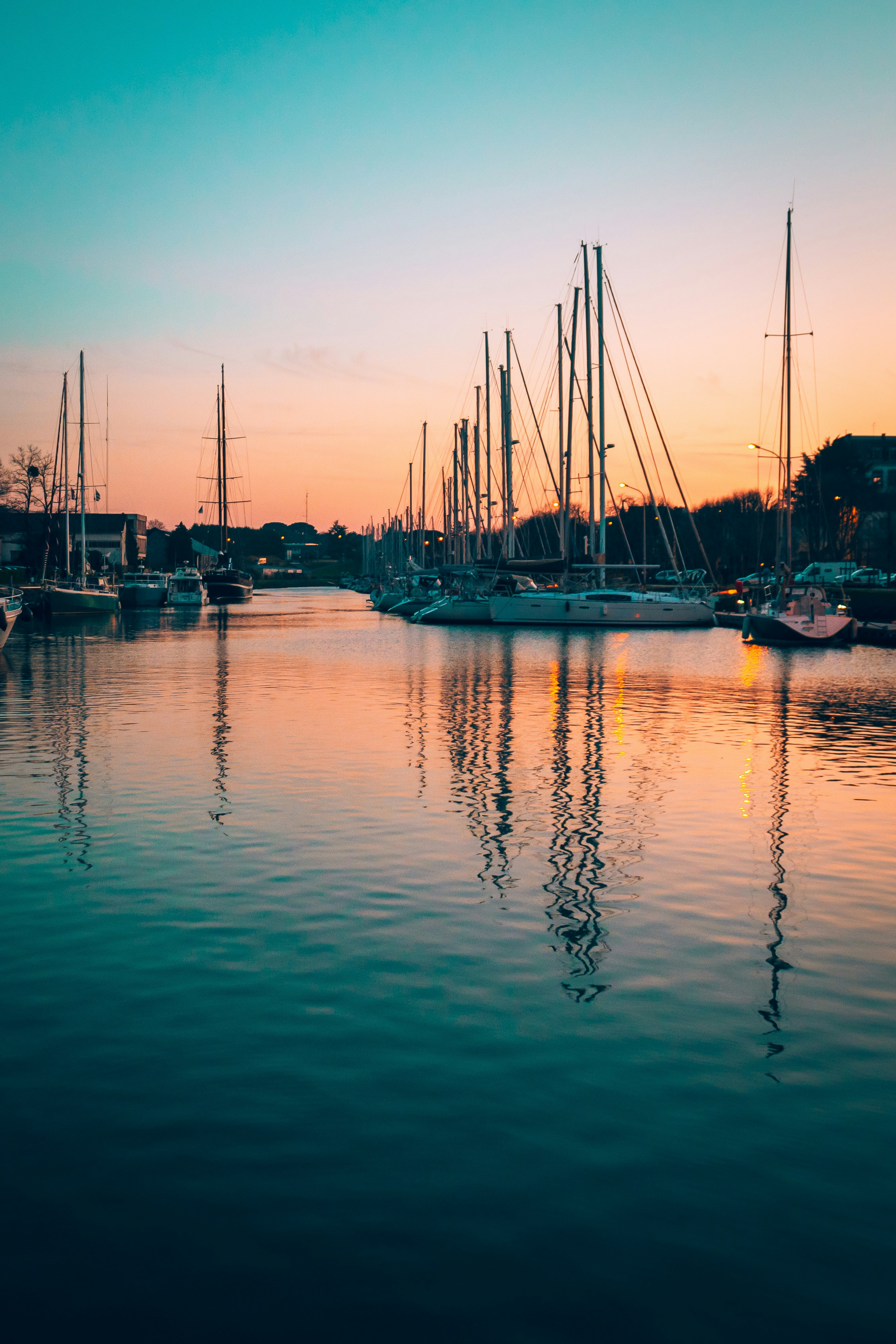 A tranquil marina scene at sunset with sailboats reflected in the calm water, surrounded by a soft orange and blue sky, creating a peaceful and serene atmosphere.