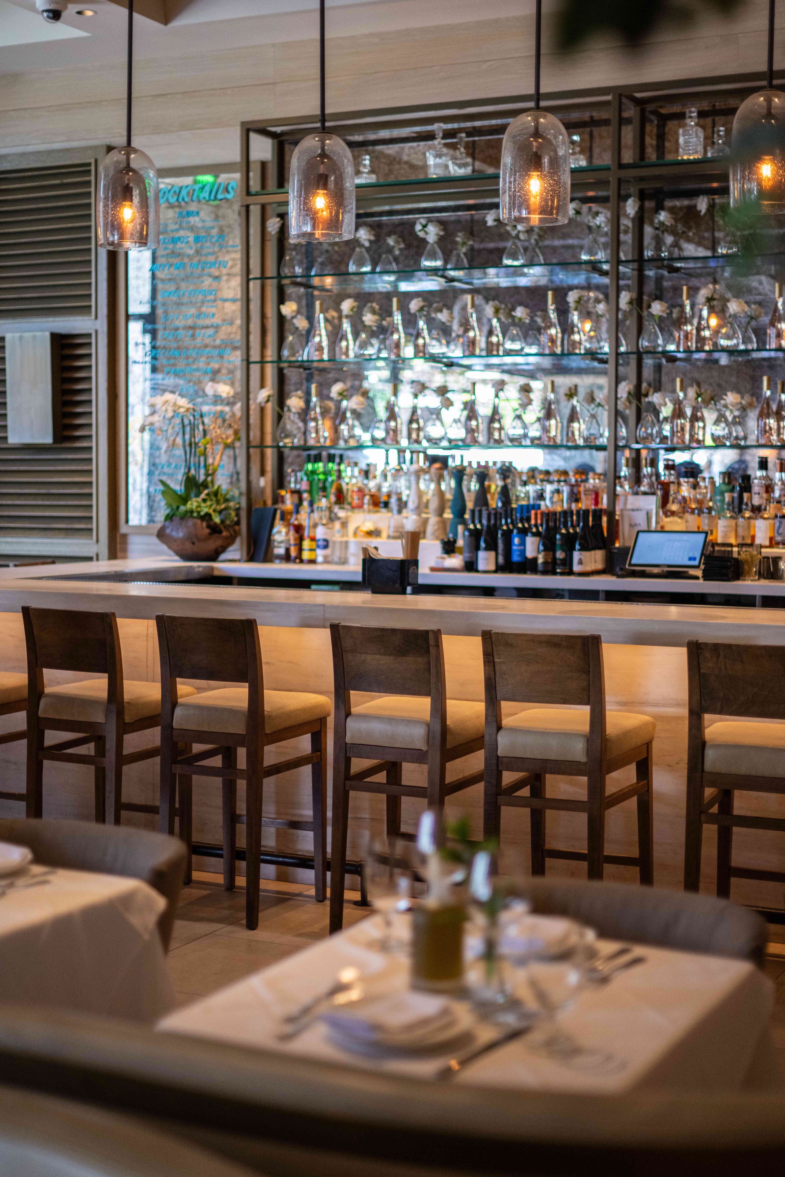 A stylish restaurant bar with tall wooden stools, elegant lighting, and a well-stocked shelf of drinks.