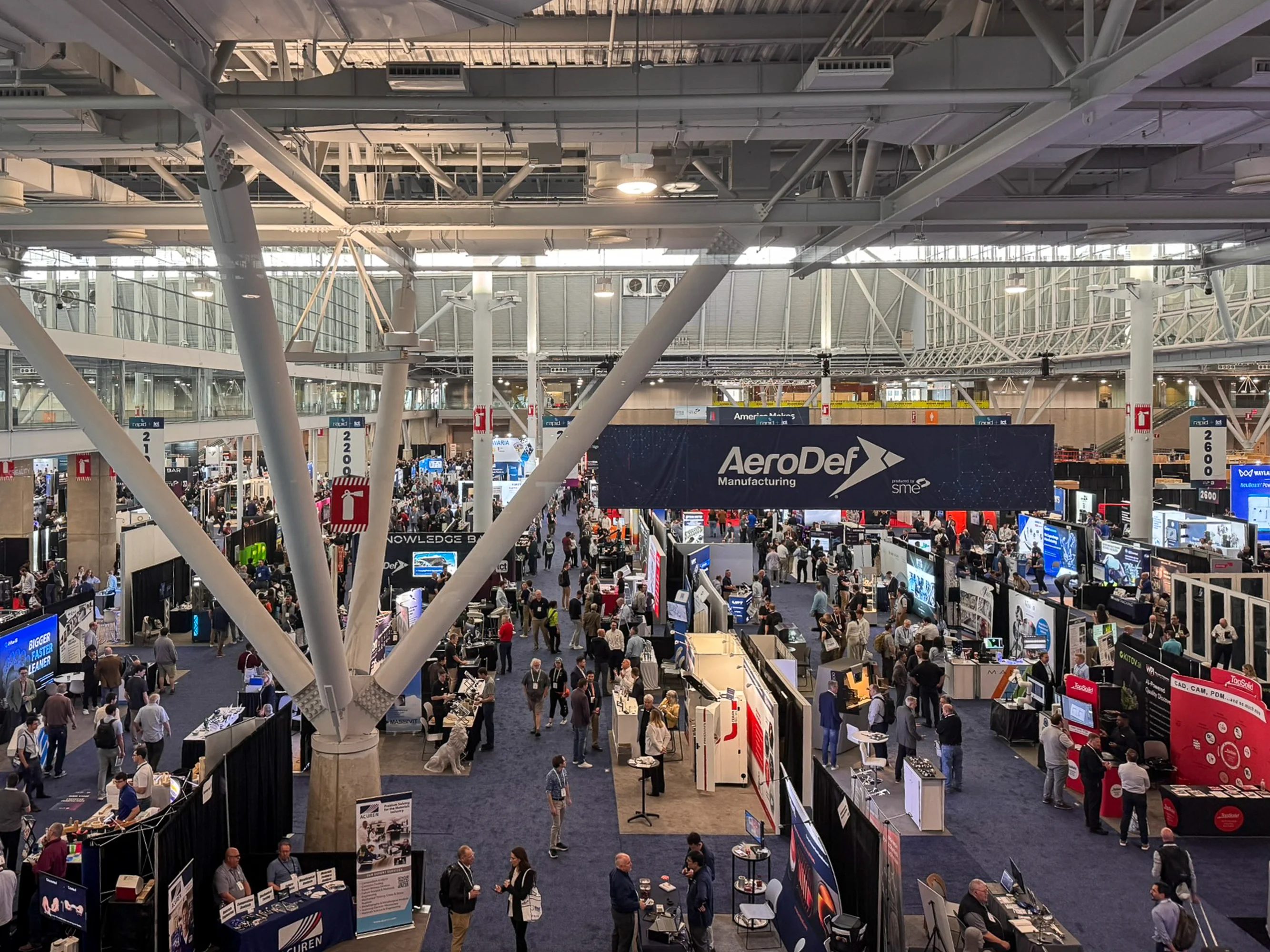 The image shows a bustling trade show floor at a large industrial convention RAPID TCT 2026, with numerous booths and attendees networking under a prominent sign for "AeroDef Manufacturing," illustrating the dynamic atmosphere of a professional industry gathering related to additive manufacturing and technology.