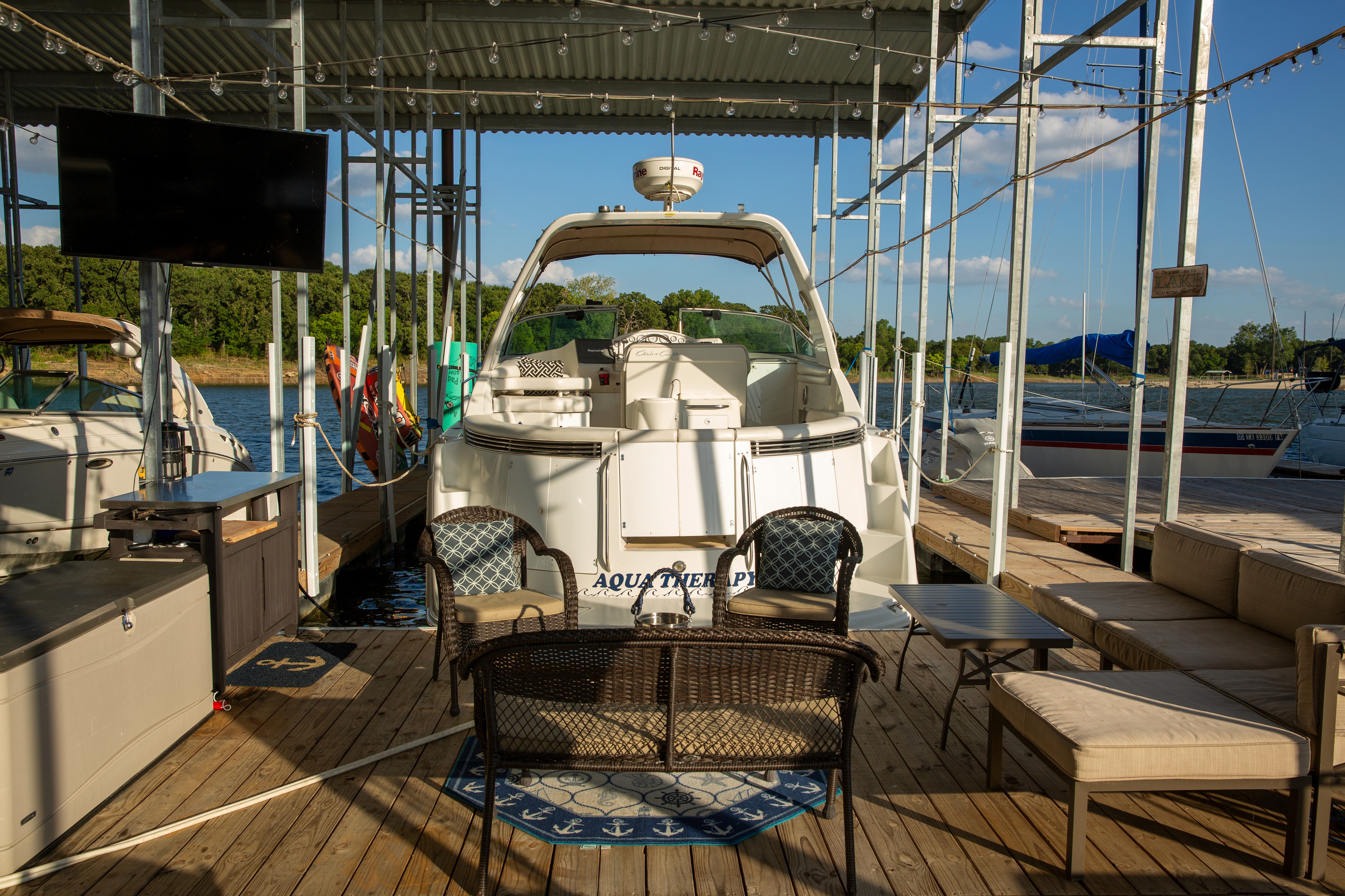 A white motorboat is docked under a shaded marina, surrounded by outdoor seating and a large flat-screen TV on a wooden deck, with a scenic view of boats and greenery in the background.