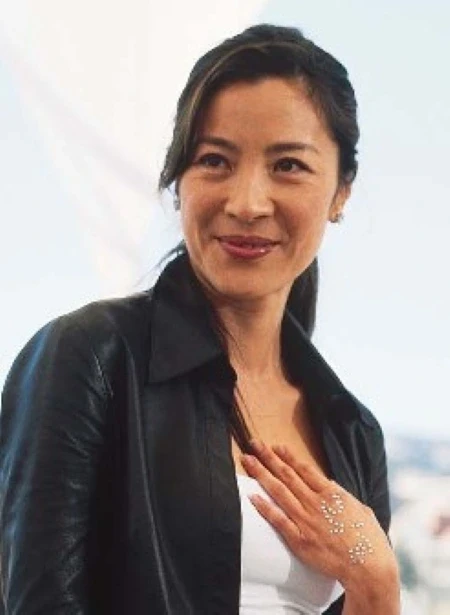 A woman with long, dark hair wearing a black leather jacket over a white top smiles and holds their hand, adorned with small metallic embellishments, near their chest against a light background.