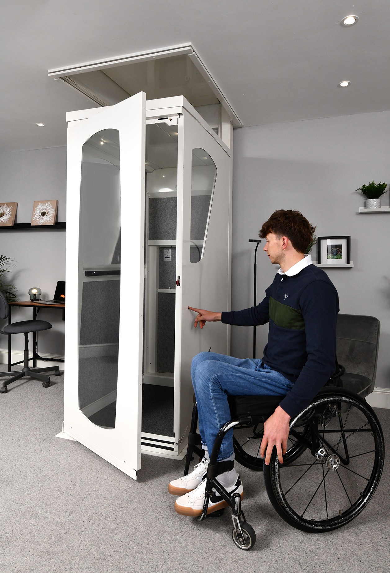 Wheelchair user operating the call button on an Elevate AVA Reach through-floor home lift with open door — white cabin with arched glass panels and grey upholstered interior installed in a modern residential living space