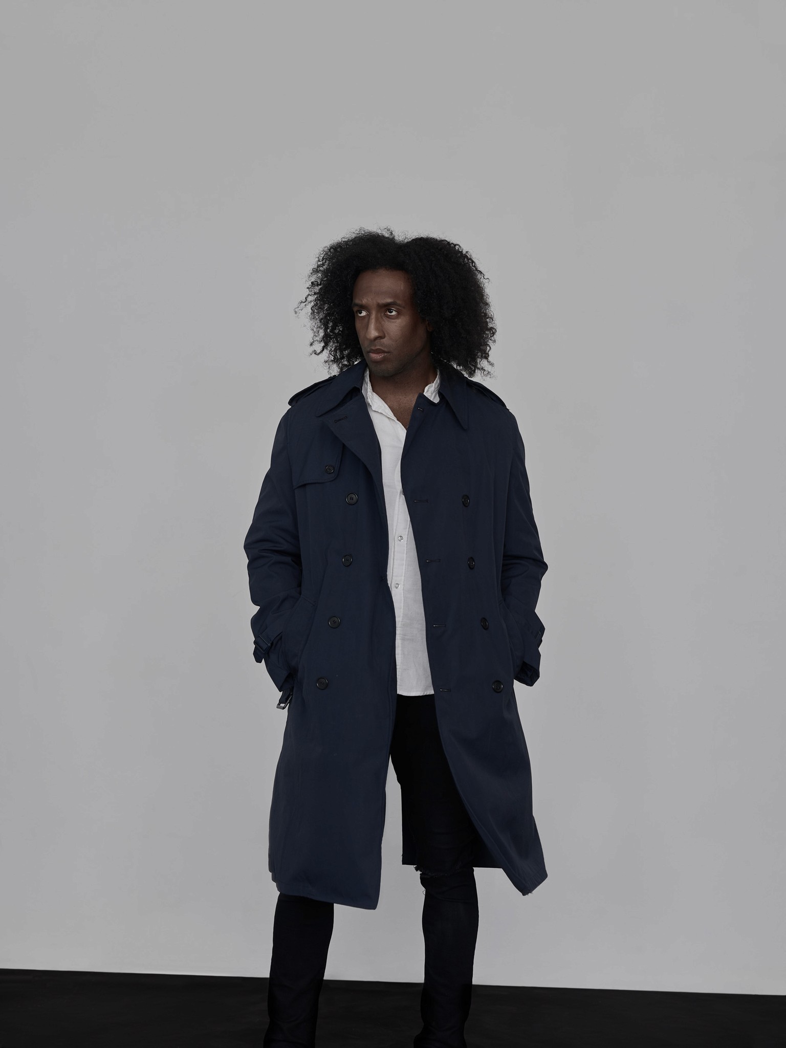 A modern photographic editorial portrait of a black man. He is wearing a trench coat and photographed in a clean graphic manner. 