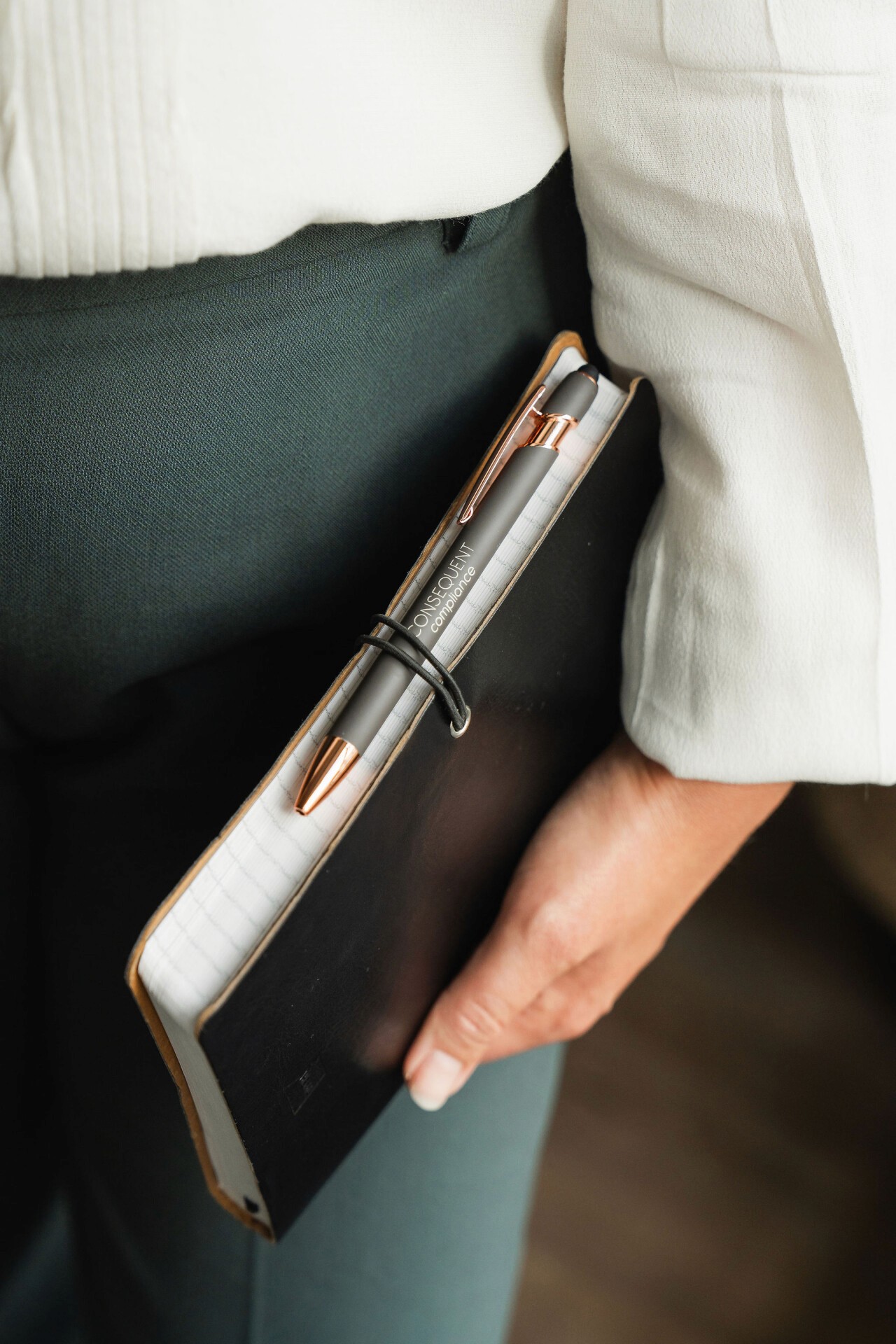 A man wearing glasses and a blazer working at a desk, writing in a notebook beside a computer monitor in a modern office.