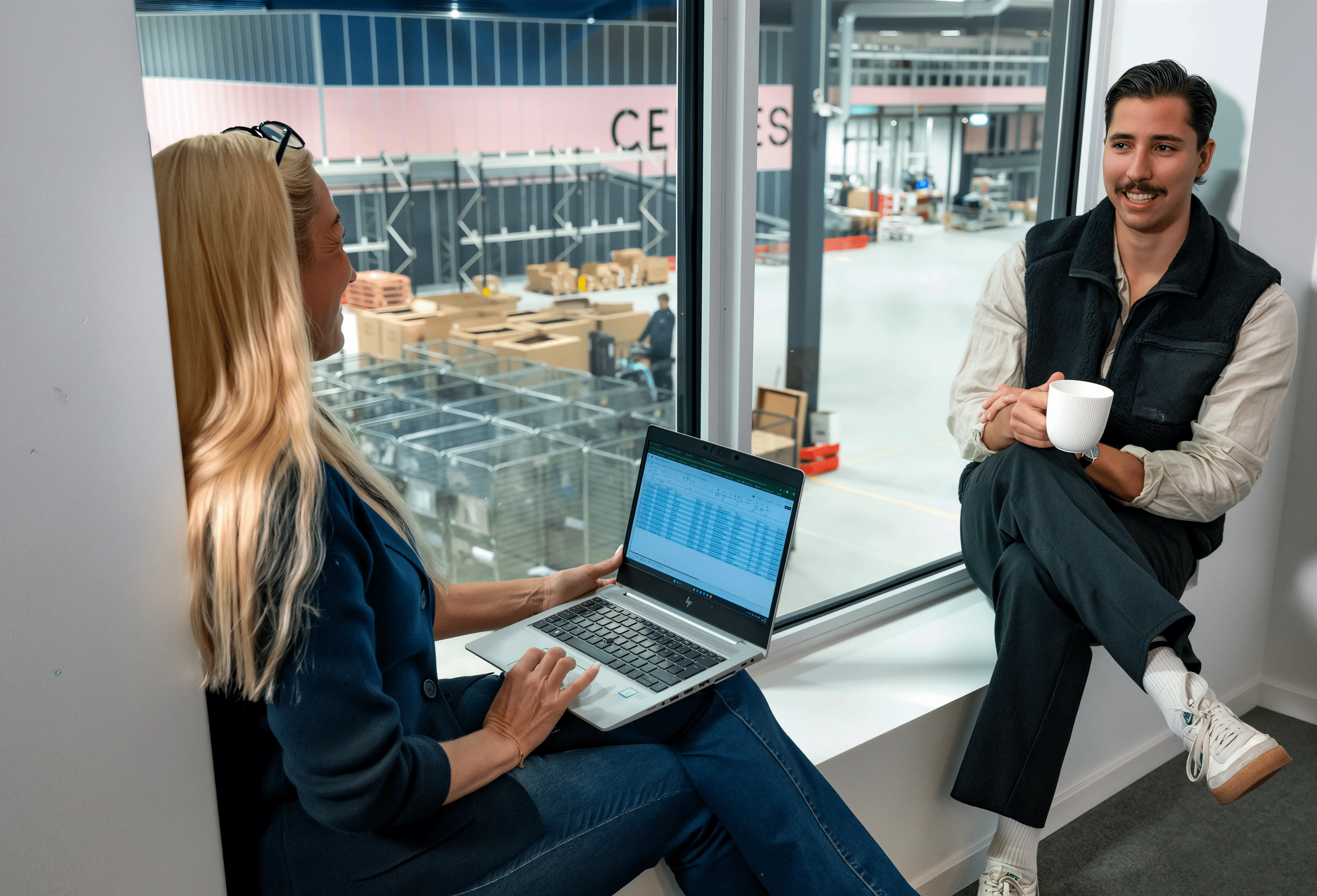 Kvinna och man diskuterar vid ett fönster. Kvinnan använder en bärbar dator. Mannen håller i en vit kaffekopp.