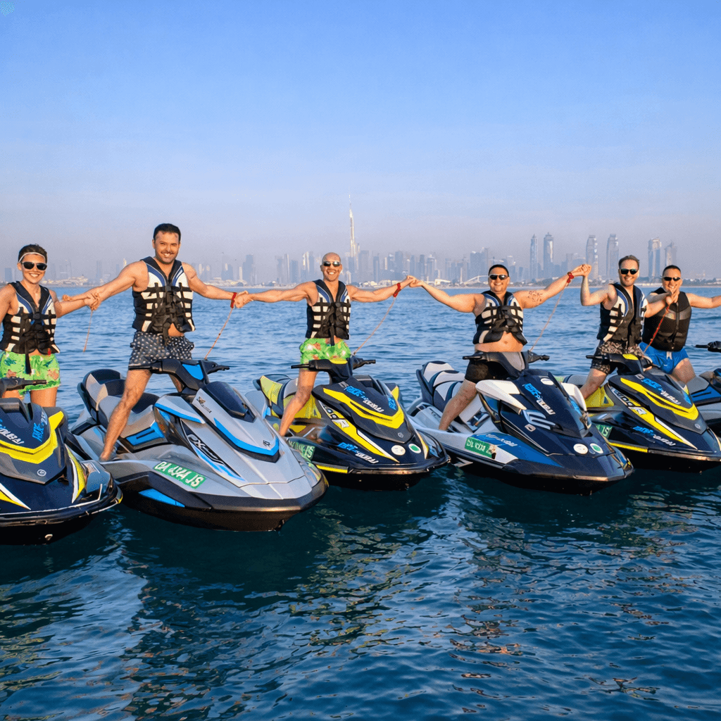A group of friends smiles and holds hands together while standing on their jet skis.