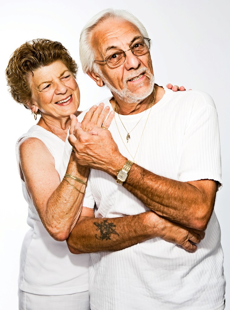 A cheerful elderly couple dressed in white embrace and smile, highlighting their loving relationship and joyful connection against a plain white background.