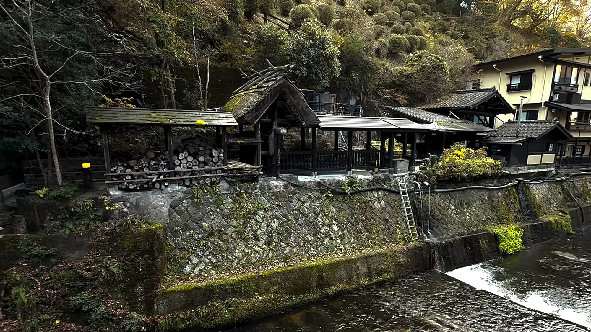 A riverside open-air bath under a timber roof beside the stone embankment at Kurokawa Onsen