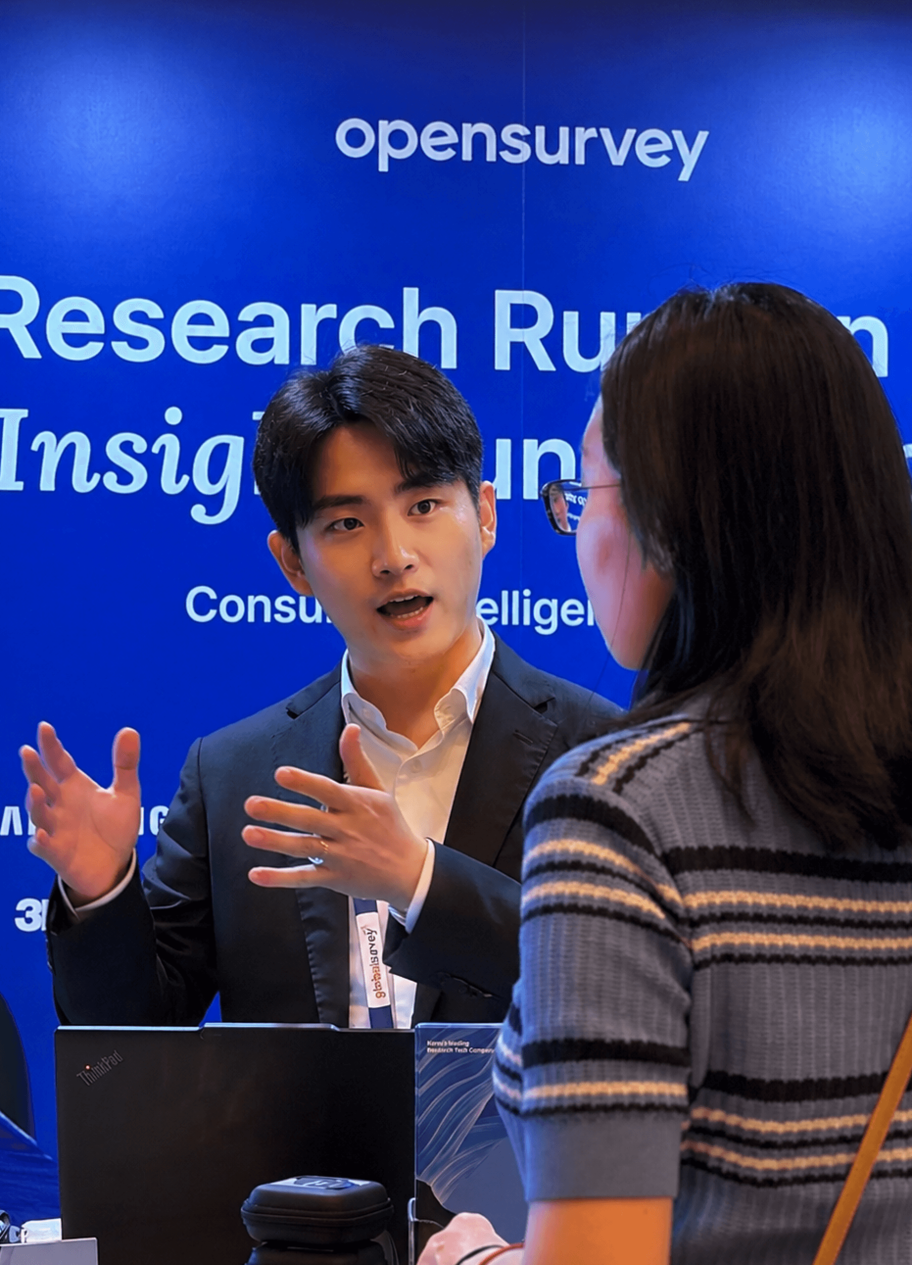 A man gestures while discussing with a woman in a business setting, with a blue background displaying research topics.