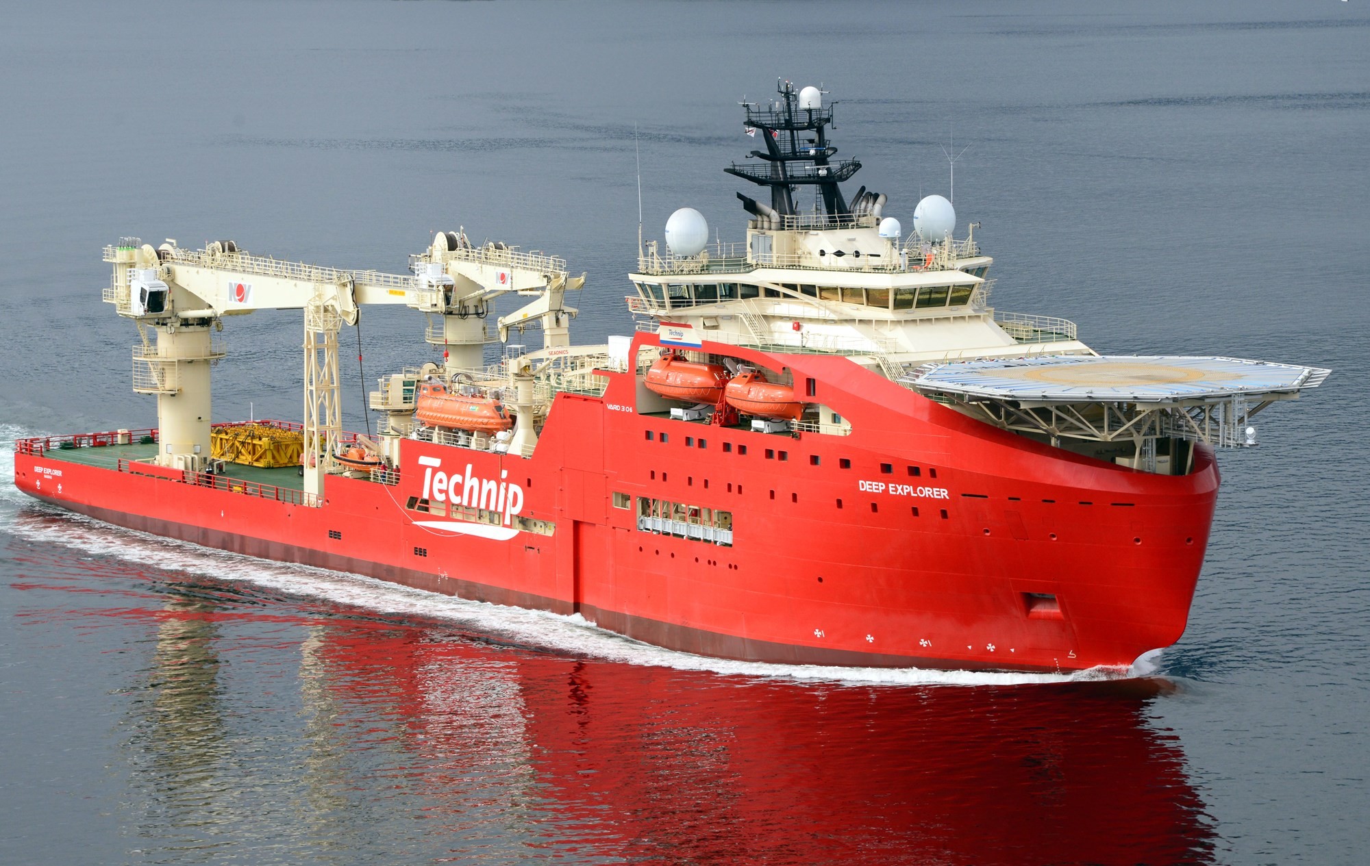 A red ship with various equipment on deck sails calmly on a body of water under clear skies.
