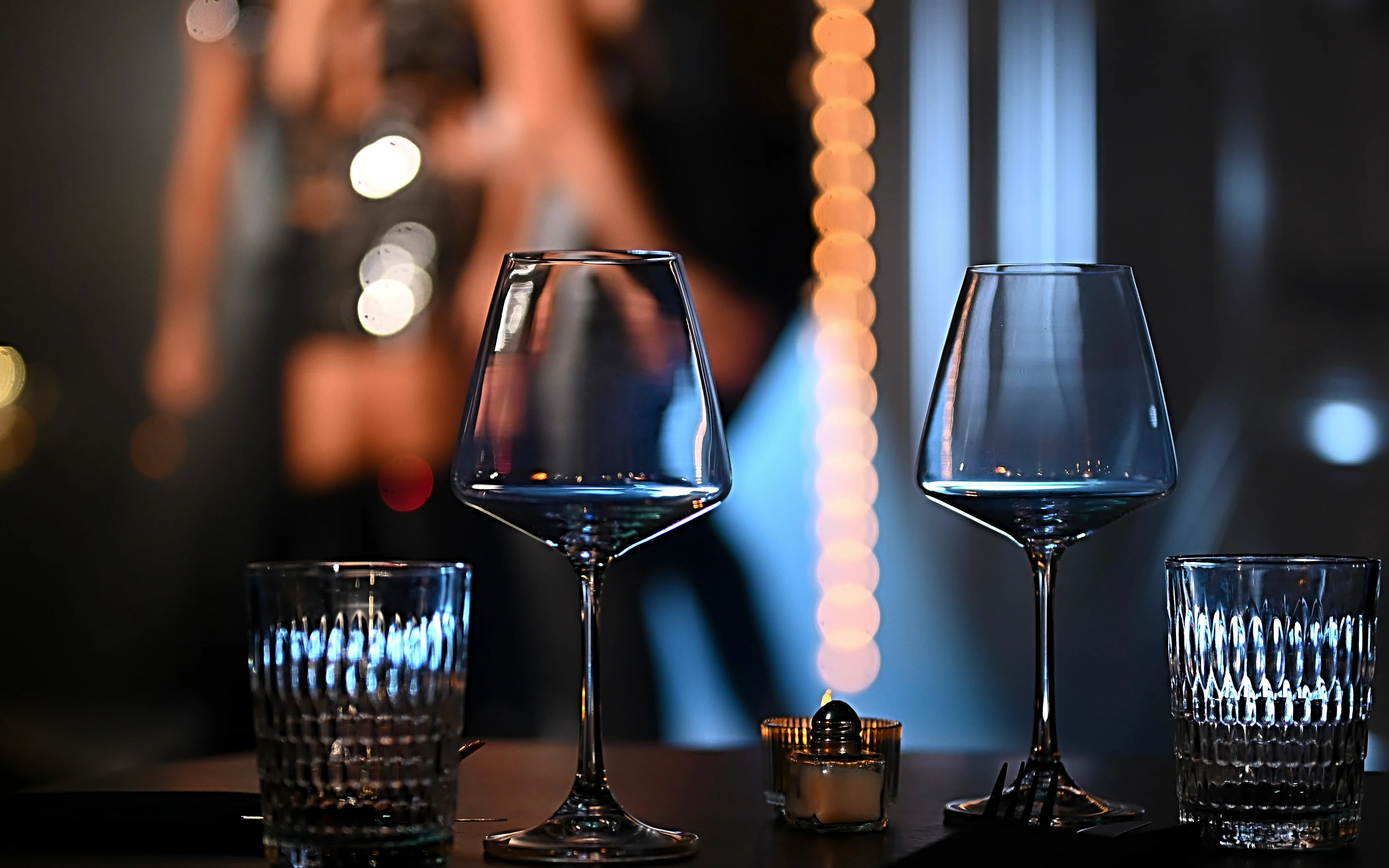 Close-up of elegant wine and water glasses resting on a dimly lit cabaret table, with warm blurred string lights in the background creating an intimate night atmosphere.