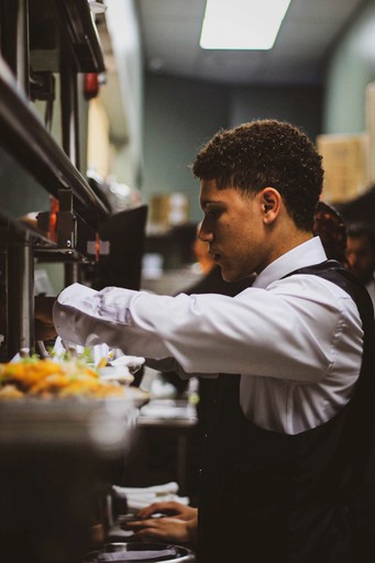 A young man in a white shirt and black vest prepares food in a commercial kitchen.