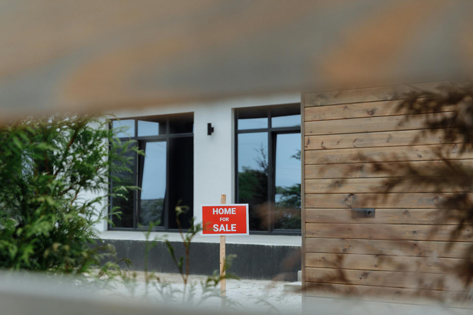 Modern house with timber cladding and large windows with a red Home For Sale sign in the front garden.