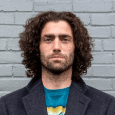 A person with curly brown hair stands against a gray brick wall, wearing a teal graphic T-shirt under a dark coat, capturing a casual and stylish urban look.