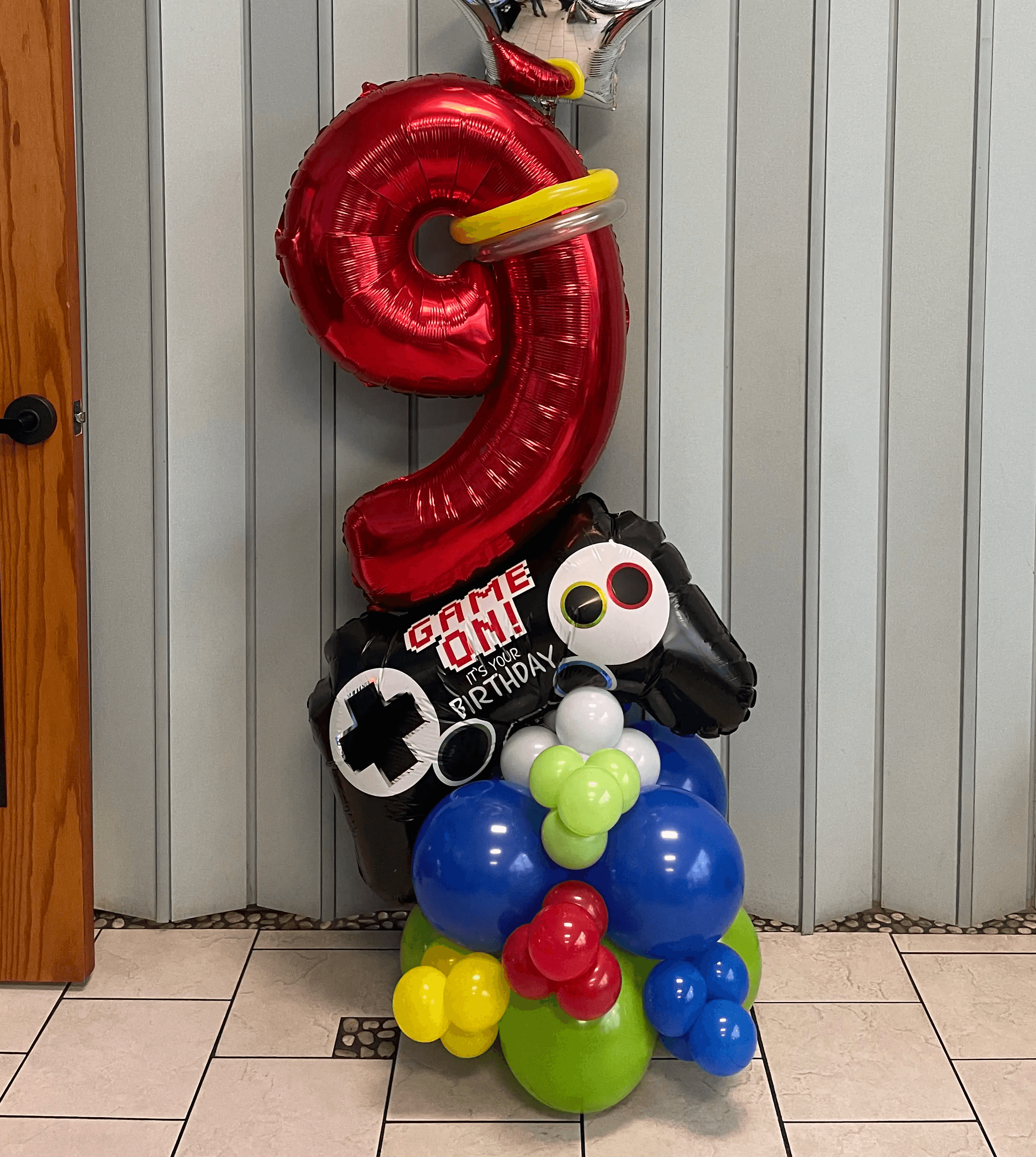 Gamer-themed birthday balloon display featuring a large red number “9” balloon, a “Game On” controller design, and bright, playful balloon clusters in blue, green, yellow, and red. Perfect for young gaming enthusiasts. Susa’s Sweets & Balloons.