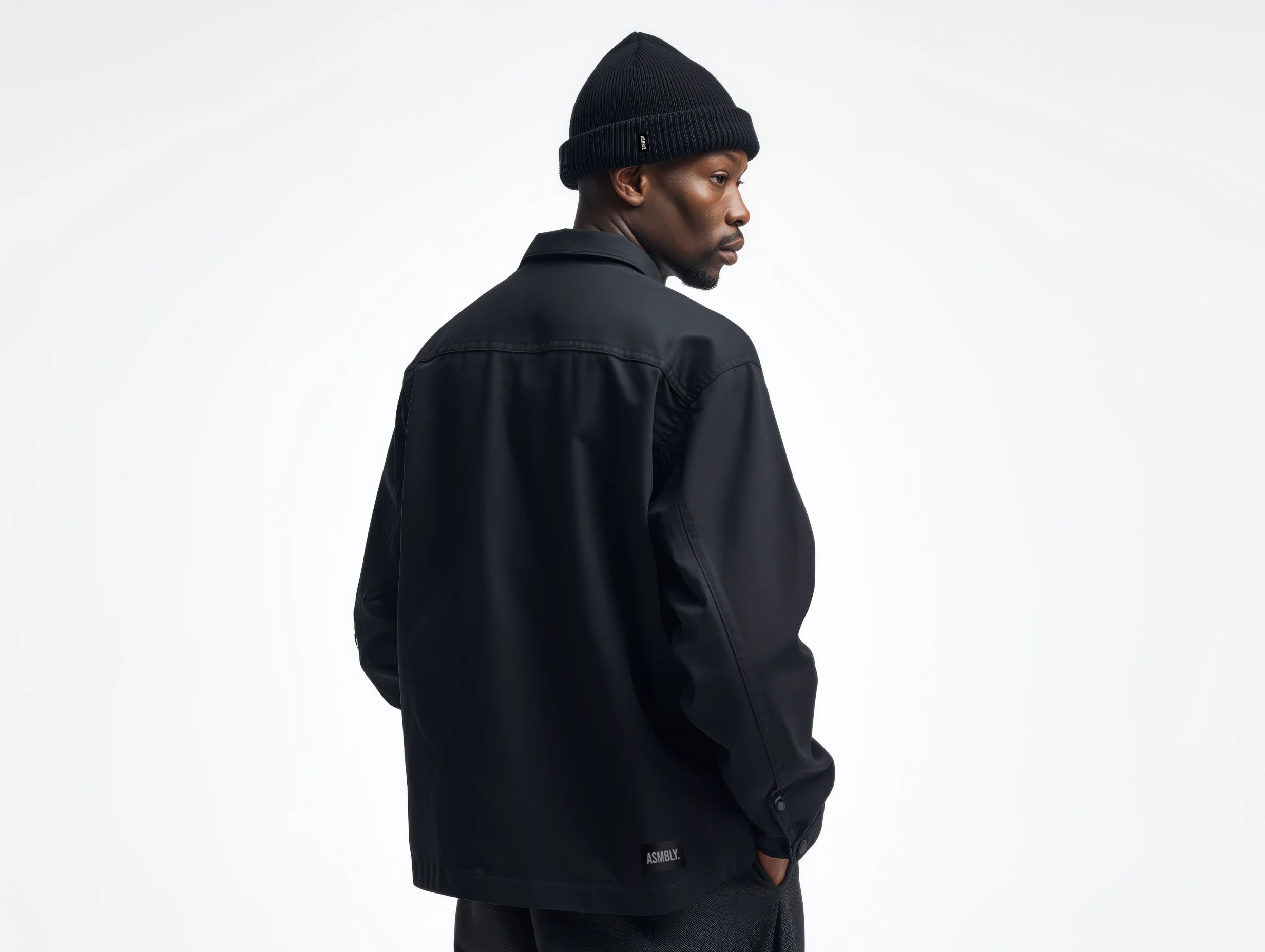 Studio portrait of a model wearing a dark jacket and beanie from Assembly fashion label, viewed from behind.