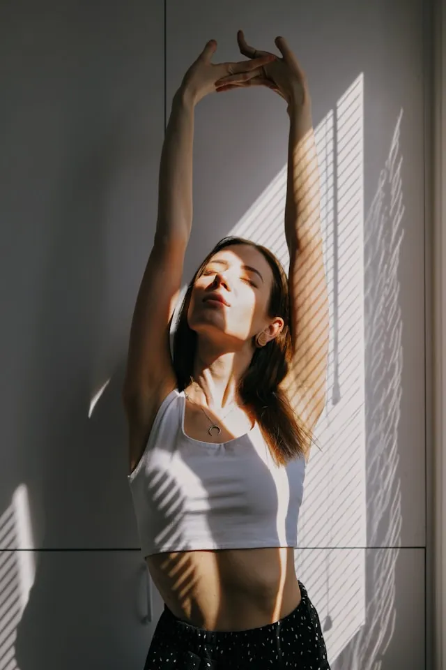 Woman stretching in warm morning sunlight by a window, eyes closed, promoting calm and daily reset.