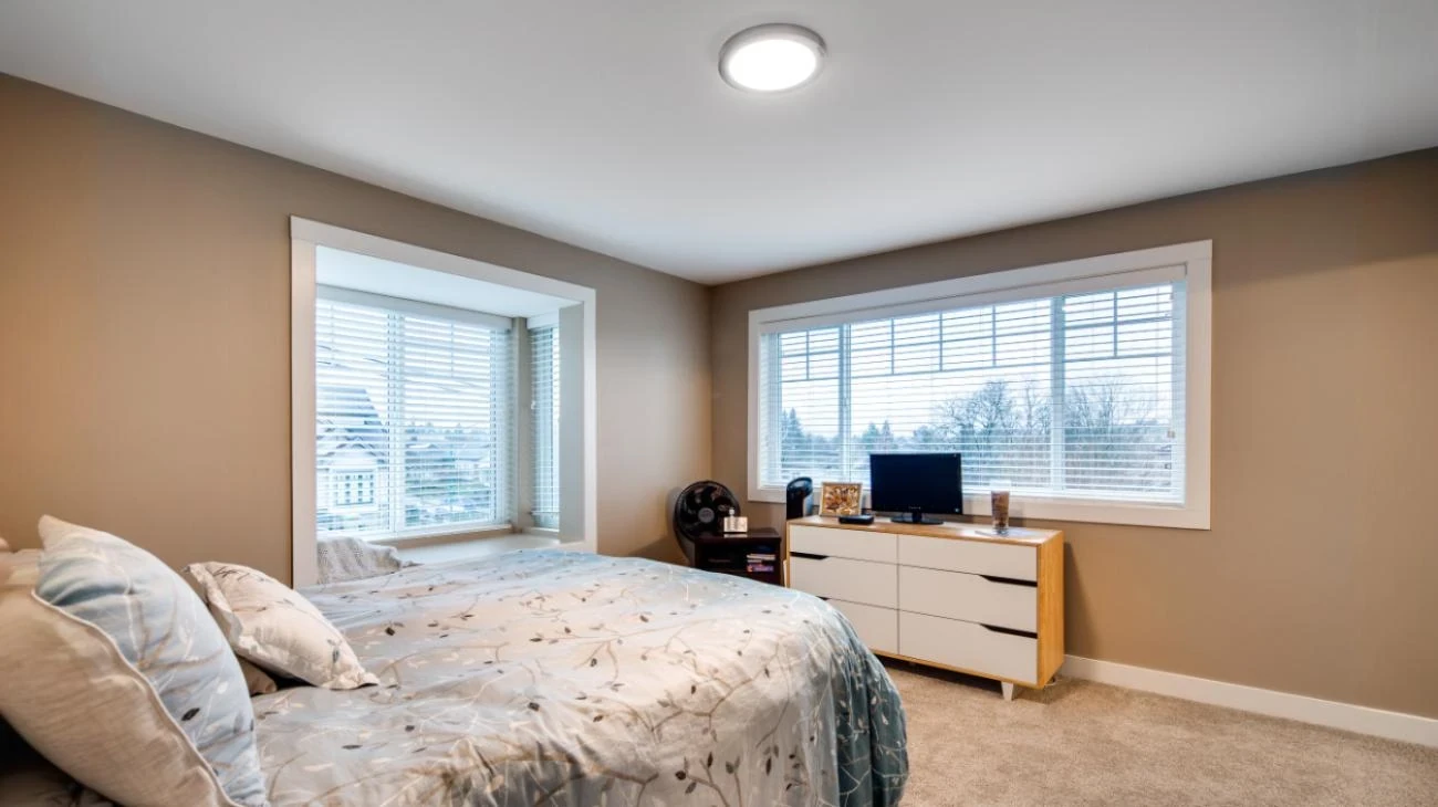 Peaceful master bedroom in custom Burnaby home with warm taupe accent walls, contemporary wood-tone and white dresser, oversized windows with privacy blinds, carpet flooring, and serene neutral palette