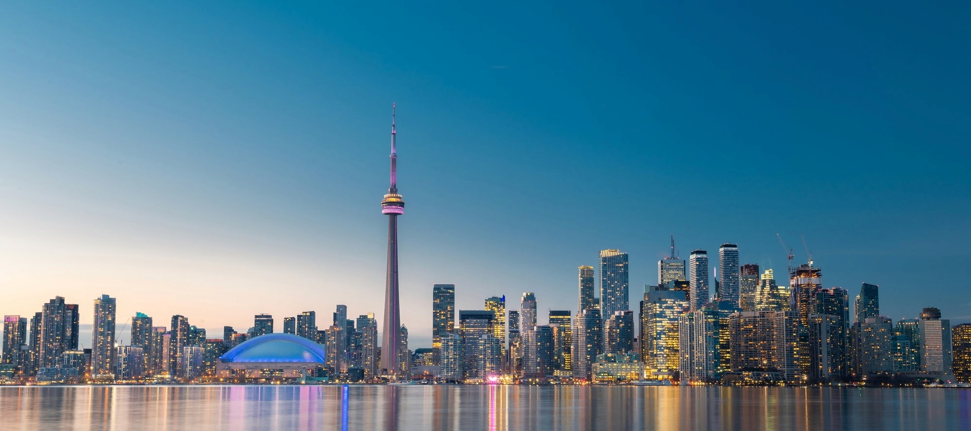 Toronto skyline at dusk with the illuminated CN Tower and city skyscrapers reflected in the calm lake water.