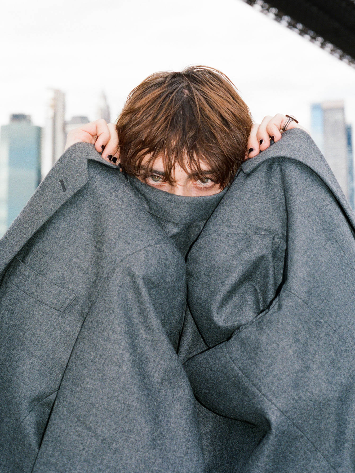 A person with short brown hair partially hides their face behind a gray coat, with urban skyscrapers and a cloudy sky visible in the background.