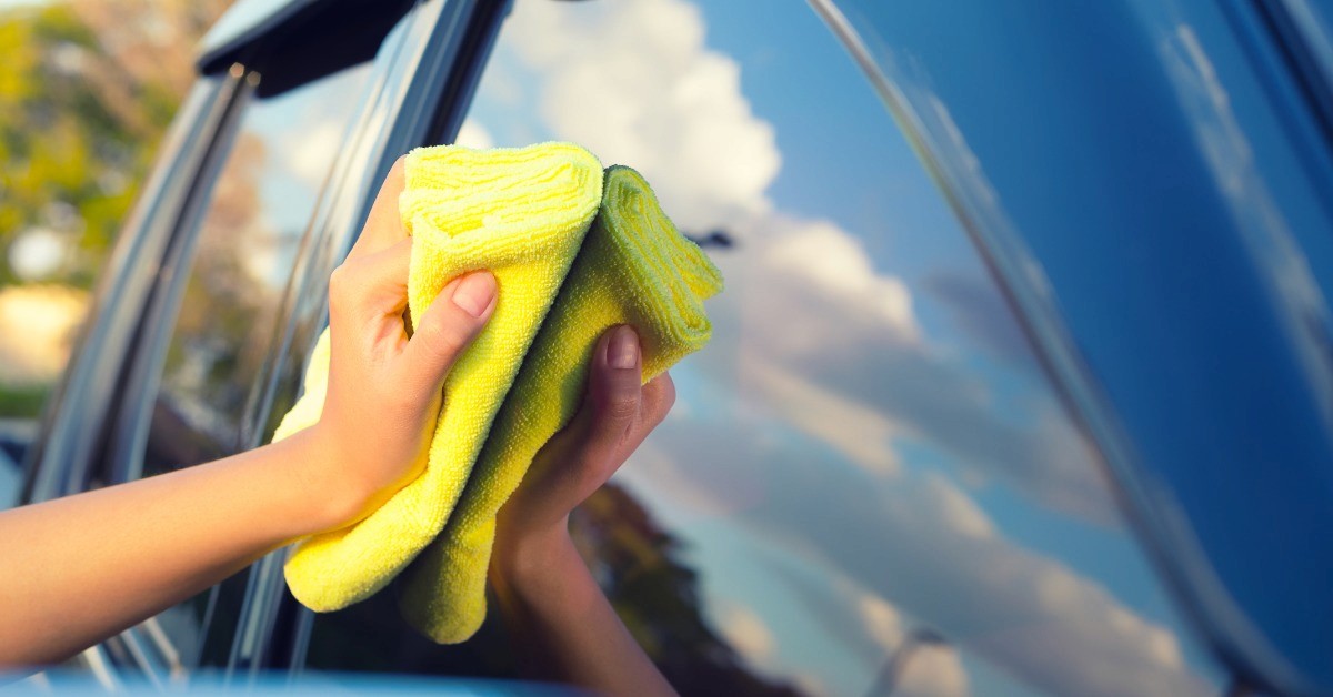 A hand holds a yellow microfiber cloth to the window of a shiny blue car. The person's arm is reflected in the window.
