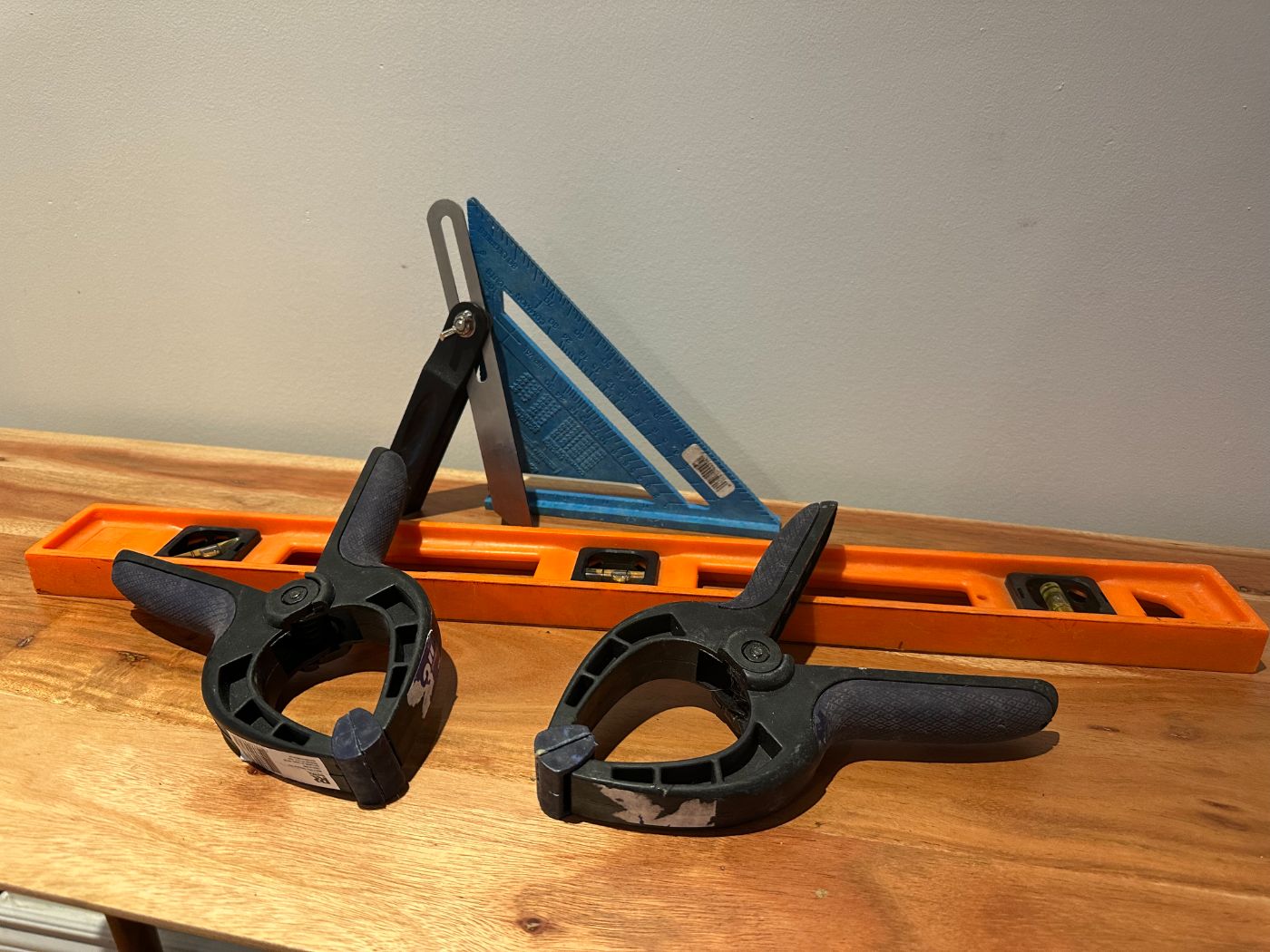 Kit d'outils de bricolage posé sur une table en bois : niveau orange, équerre bleue, fausse équerre métallique et deux serre-joints noirs