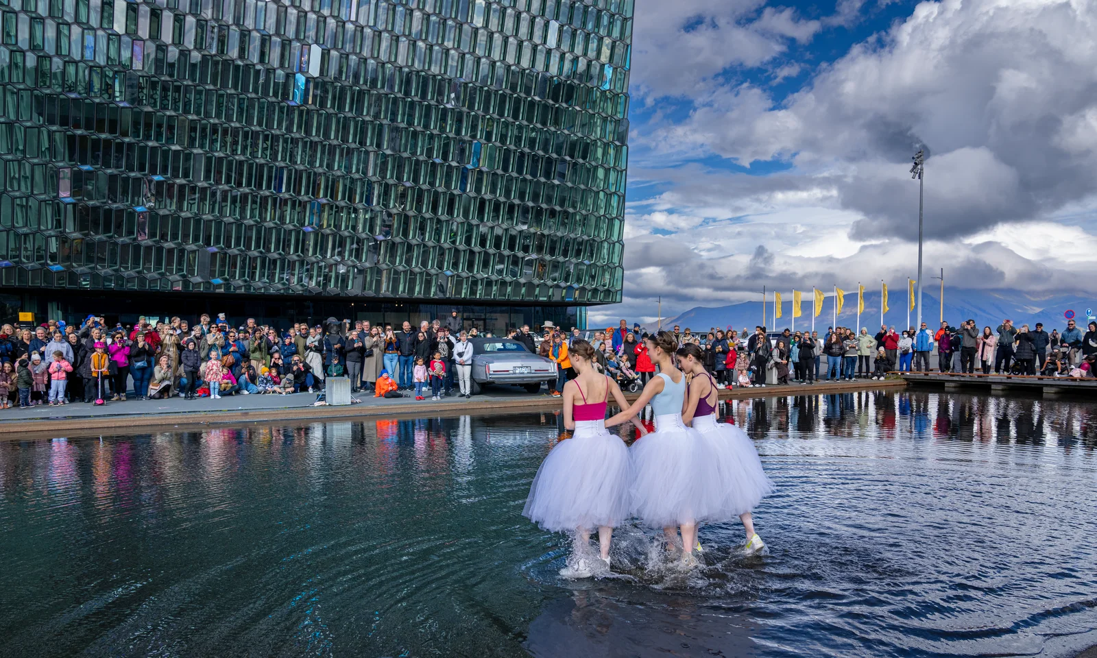 Three ballet dancers in white tutus perform a routine while wading through a shallow pool of water outside the Harpa Concert Hall during Reykjavík Culture Night. A large crowd of spectators stands along the edge watching the outdoor performance under a bright, cloudy sky.