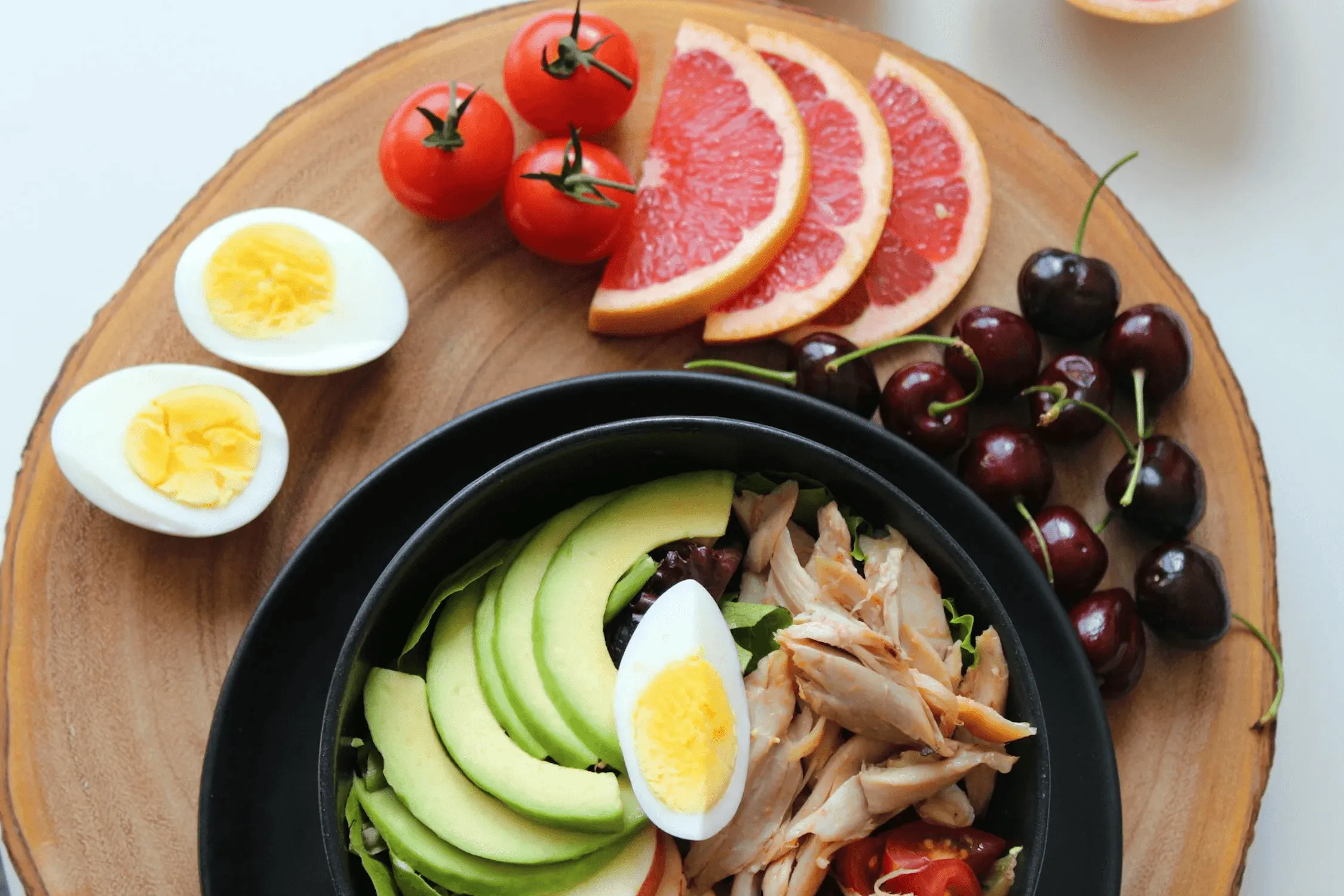 A vibrant meal featuring sliced avocado, eggs, cherry tomatoes, grapefruit, and grapes on a wooden platter.