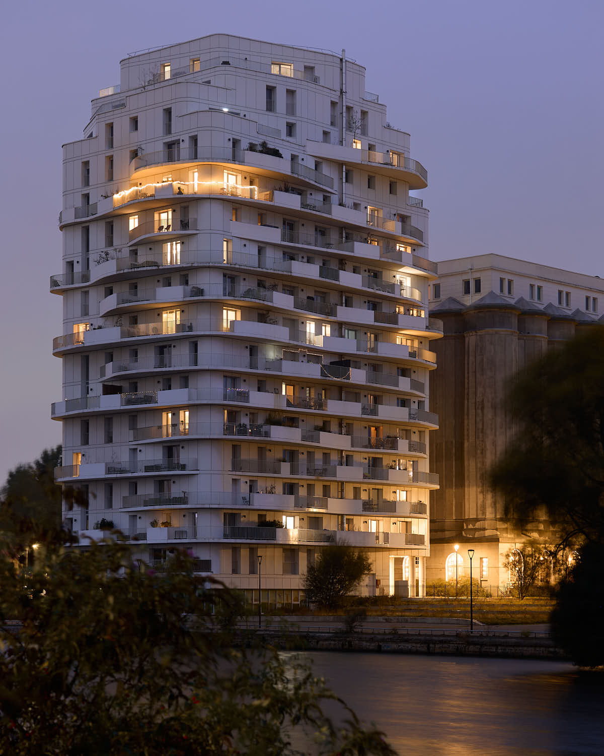 Vue au crépuscule avec un ciel légèrement violet de la Brooklyn Tower de Marquette-lez-Lille, avec quelques fenêtres éclairer de l'intérieur en jaune, photo prise de l'autre côté de la Deûle avec en arrière-plan à droite, sur les anciens silos en béton des Grands Moulins de Paris.