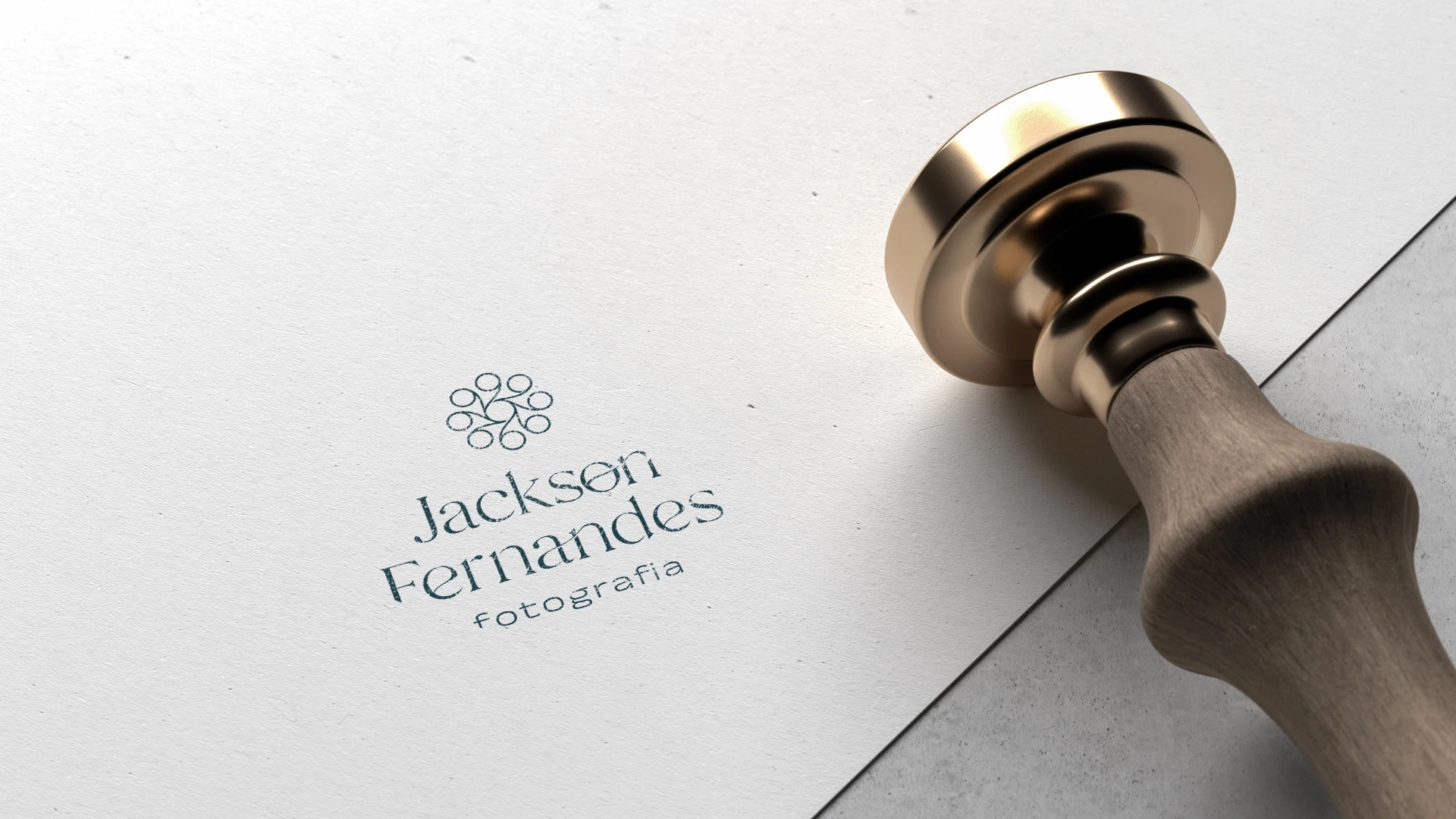 Sinete metálico dourado com cabo de madeira apoiado sobre papel texturizado claro, exibindo a marca Jackson Fernandes Fotografia gravada em relevo na superfície.