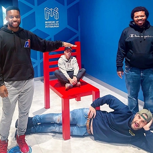 四名男子在匹兹堡幻觉博物馆的一个强迫透视展览中与一把巨大的红色椅子合影。