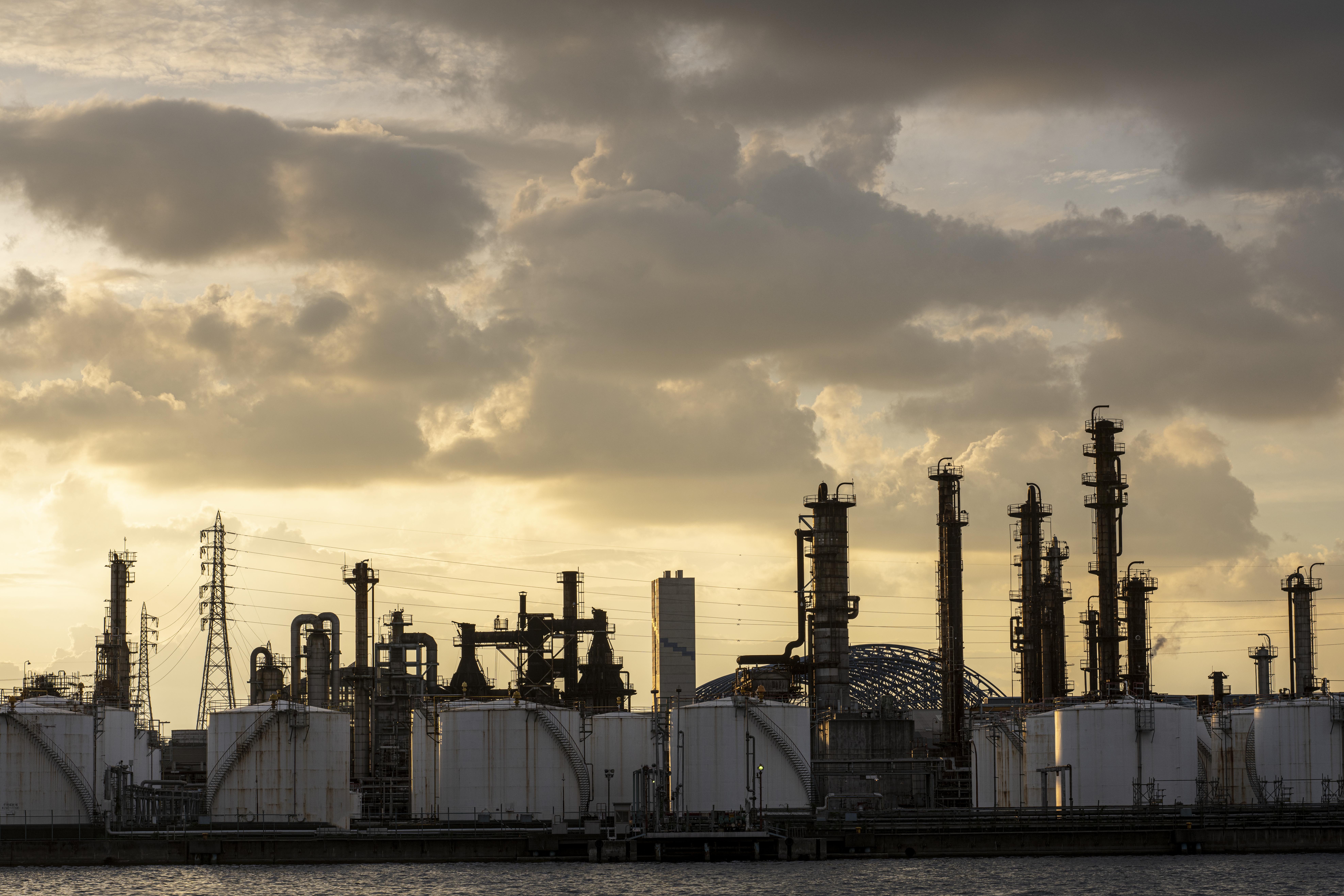 Industrial refinery silhouette at sunset, with tall chimneys and tanks under a dramatic, cloudy sky. The scene conveys a gritty, atmospheric tone.