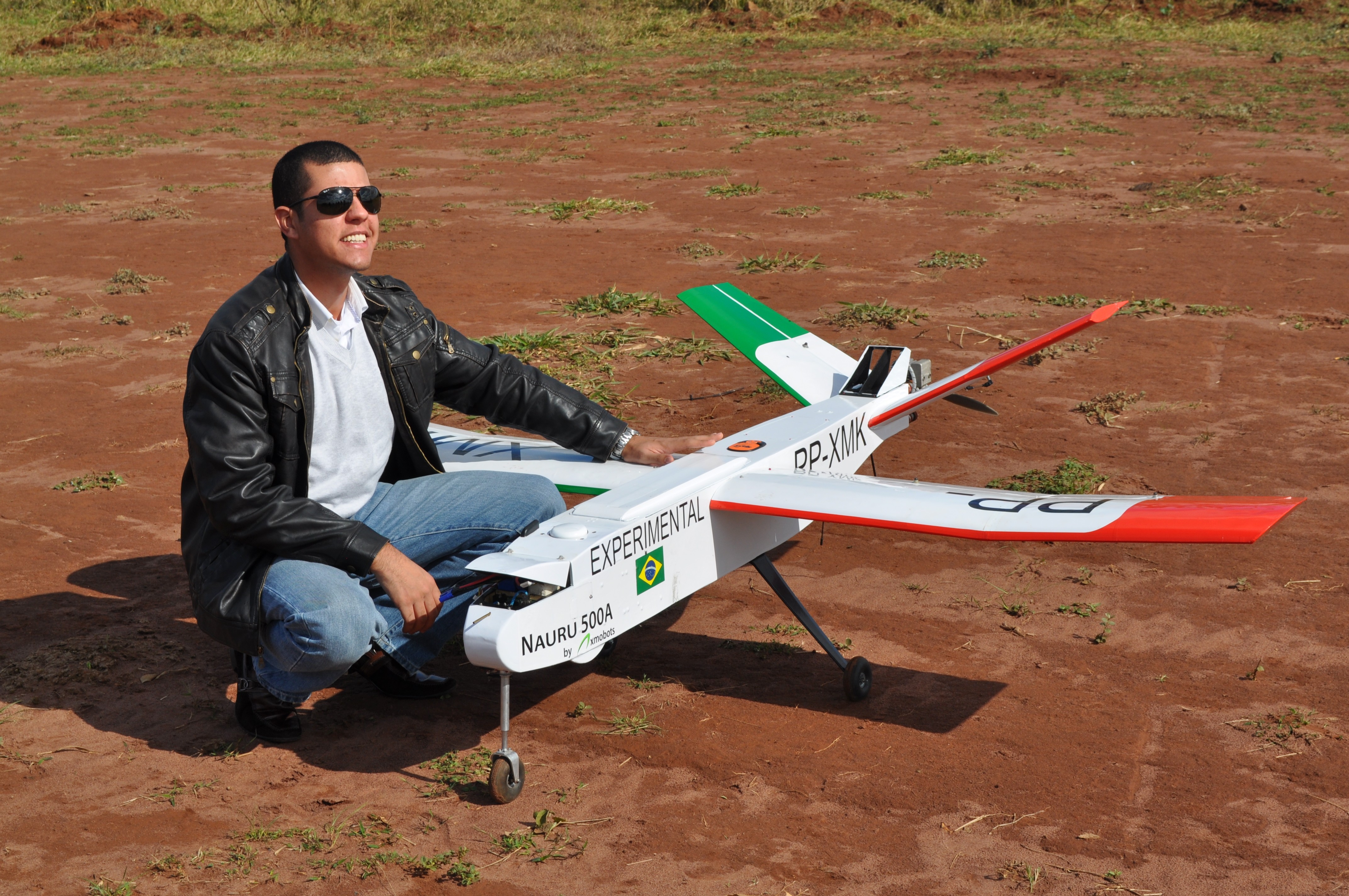 Engenheiro agachado ao lado de um drone de mapeamento aéreo experimental modelo Nauru 500A com a bandeira do Brasil em terreno de terra.