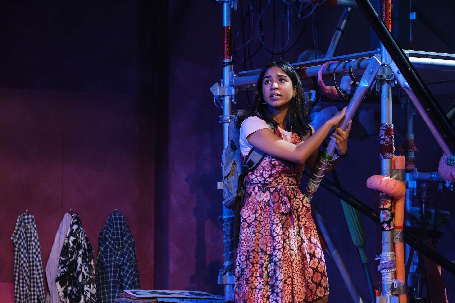 A performer in a patterned dress and backpack climbs a scaffold structure, gripping a metal pole while looking upward with urgency. The set around them is filled with cables, props, and colourful fabric.