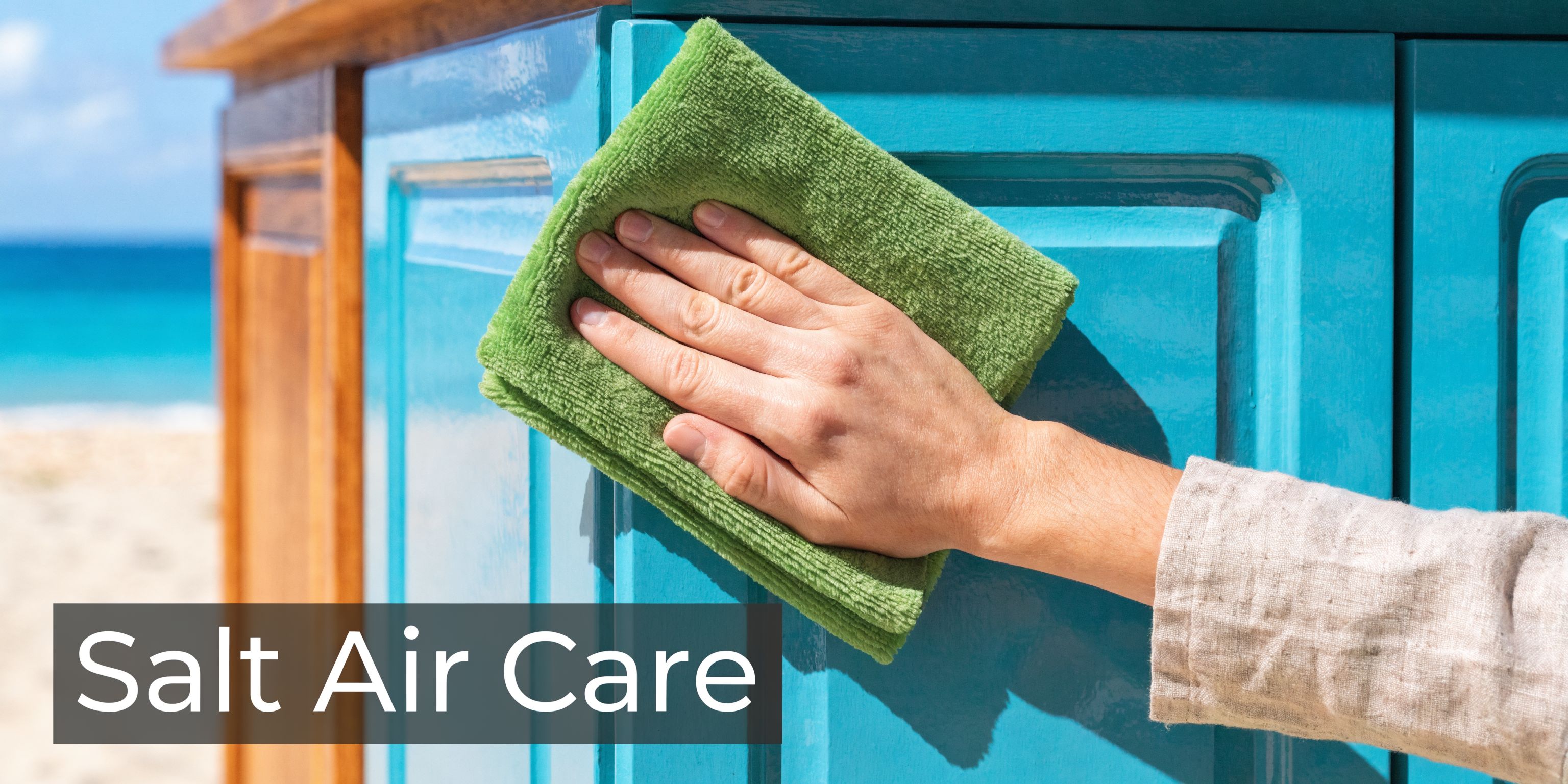 A hand wiping down blue painted kitchen cabinets with a green microfiber cloth near the beach.