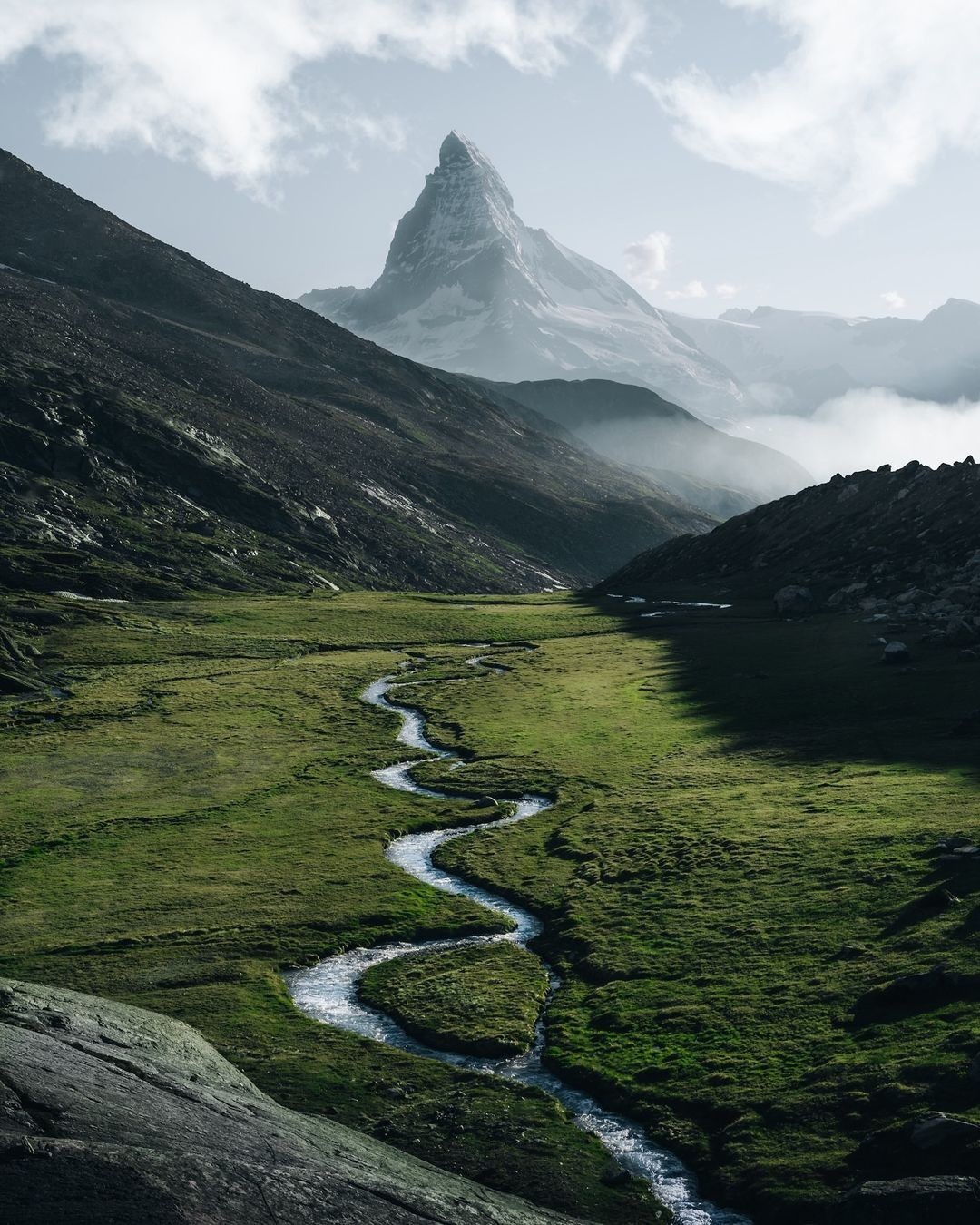 A winding river cutting through a wide alpine meadow, leading toward a sharp snow-capped mountain peak surrounded by dramatic clouds and mist.