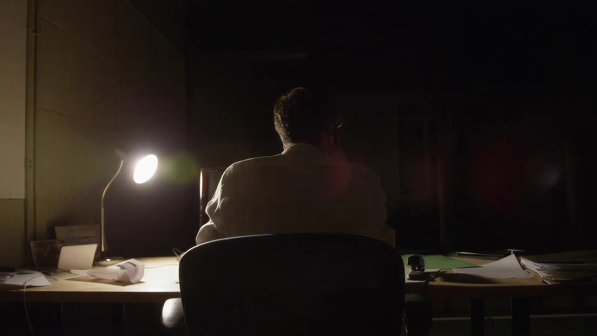 Person silhouetted from behind, sitting alone at a desk lit by a single lamp in a dark room.