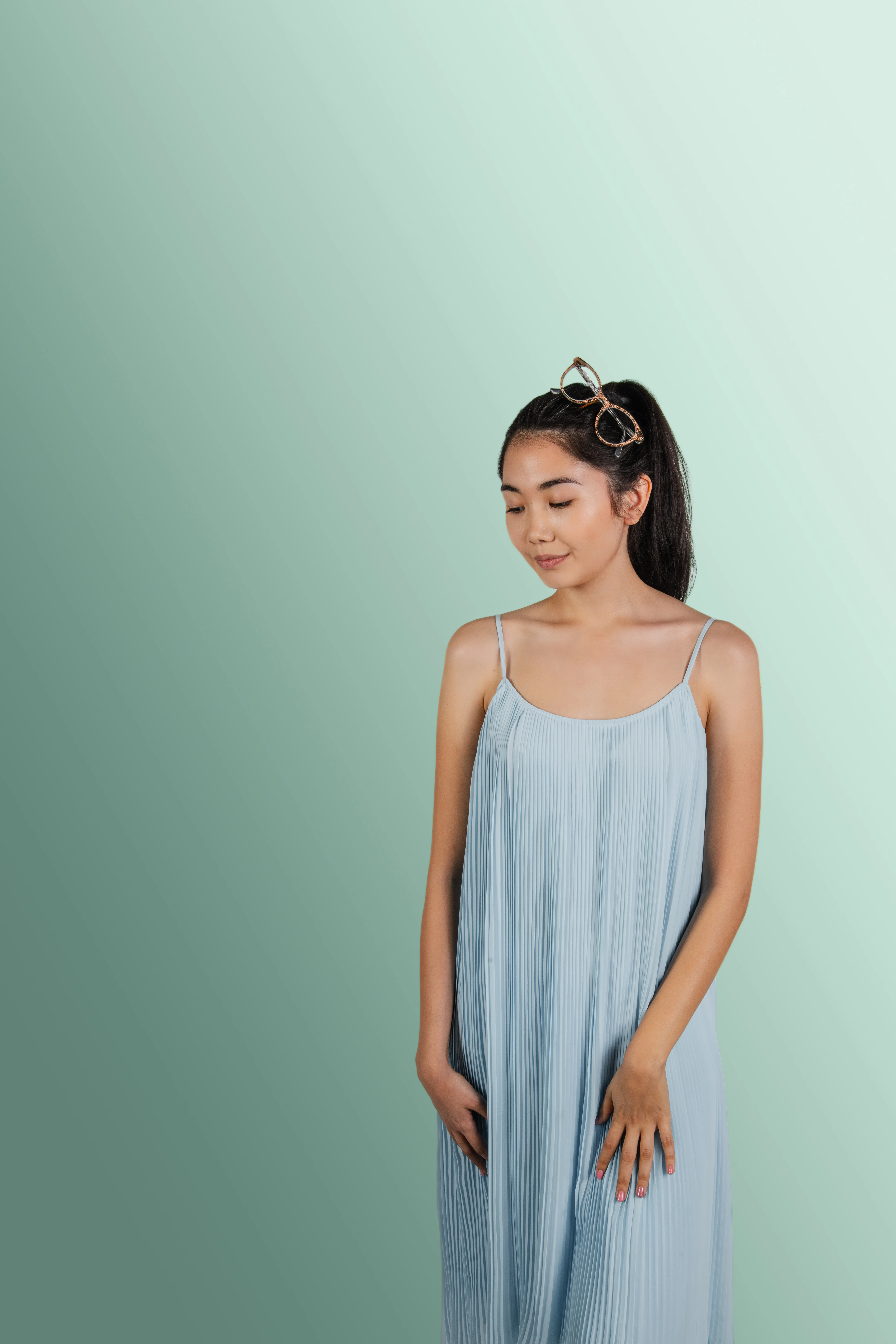 Studio portrait of an East Asian female model wearing a light blue pleated slip dress and tortoise-shell glasses in her hair. Looking down, hands on hips, set against a smooth green gradient background. High-key commercial fashion photography.