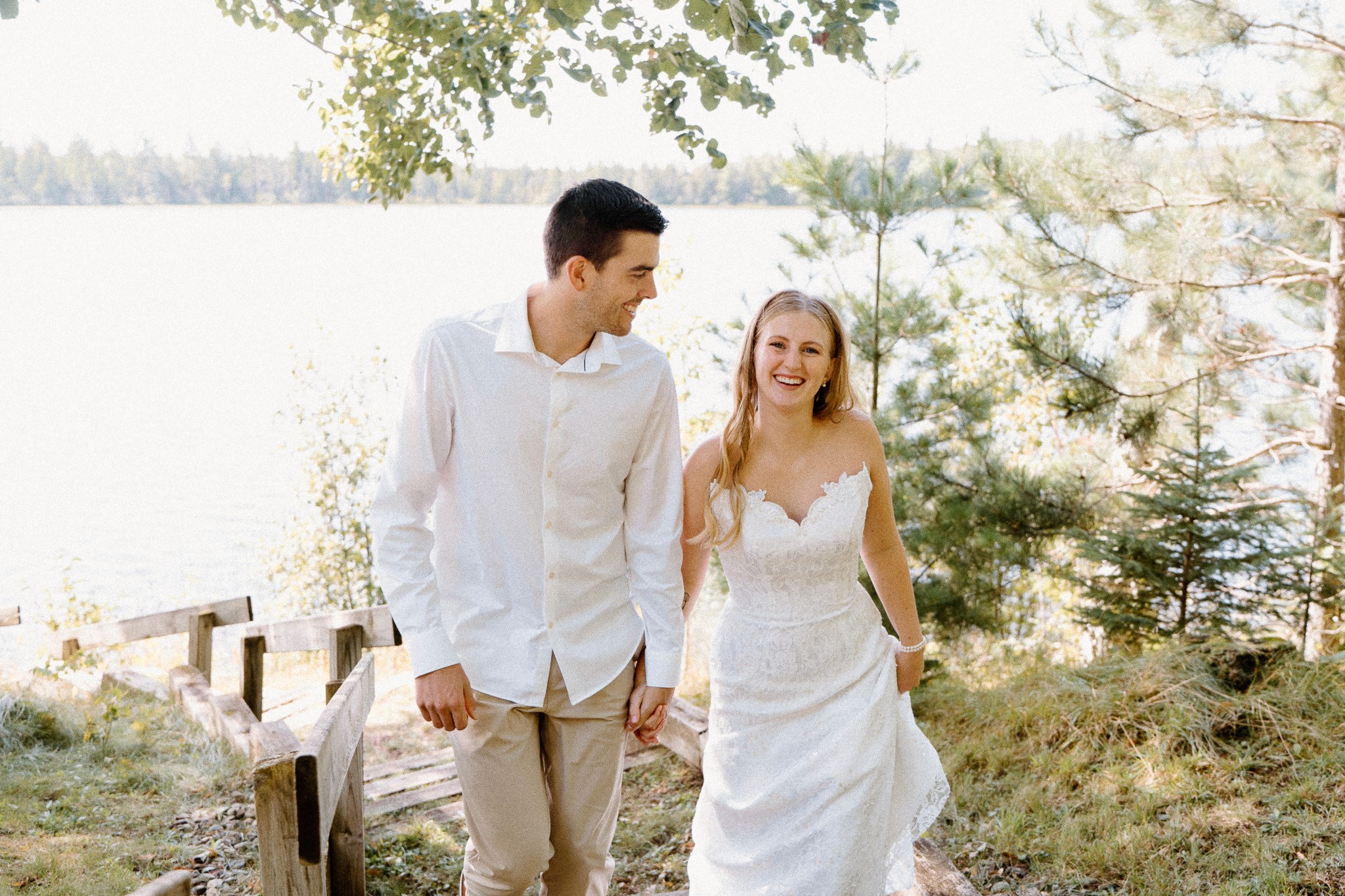 A couple walks hand-in-hand near a serene body of water, surrounded by greenery, dressed casually in light clothing.
