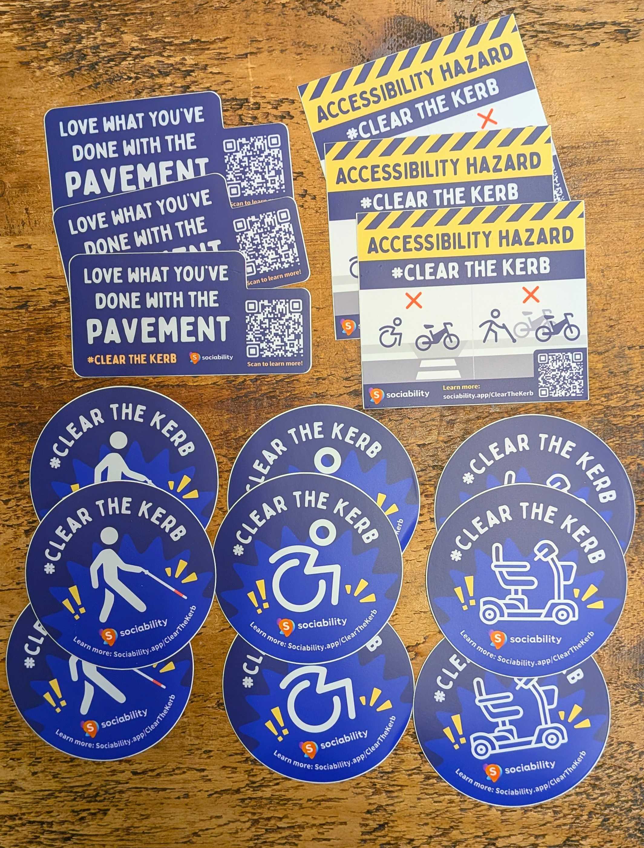 Multiple stickers advertising the Clear The Kerb campaign. One design reads 'Love what you've done with the pavement'. Another reads 'Accessibility Hazard' and looks like a parking ticket. There are three circular designs with either a cane user, wheelchair user or mobility scooter.