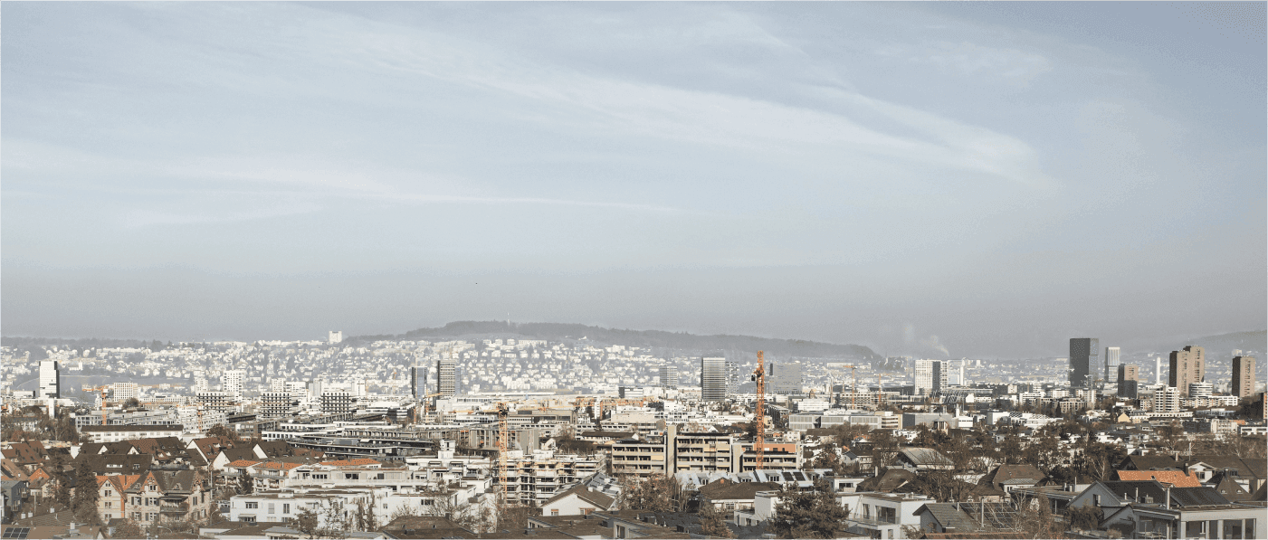 Weitläufige Panoramaansicht über Zürich mit Blick auf Wohnquartiere, Baukräne und die urbane Skyline vor leicht dunstigem Himmel.