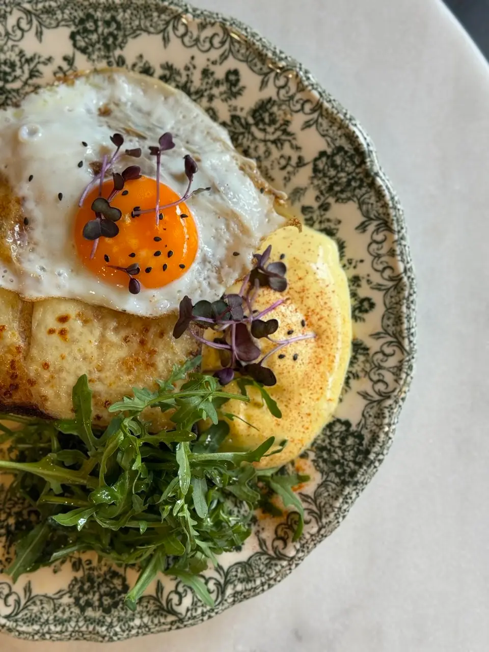 - Two artfully plated brunch dishes on speckled ceramic plates featuring eggs, greens, and colorful garnishes on a dark table