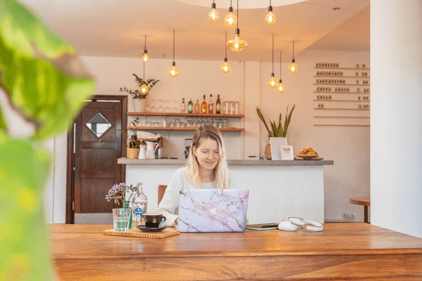 Una mujer trabaja frente a su portátil. Mesa de madera, atmósfera cálida de cafetería. Al fondo, luces colgantes y la barra de café.