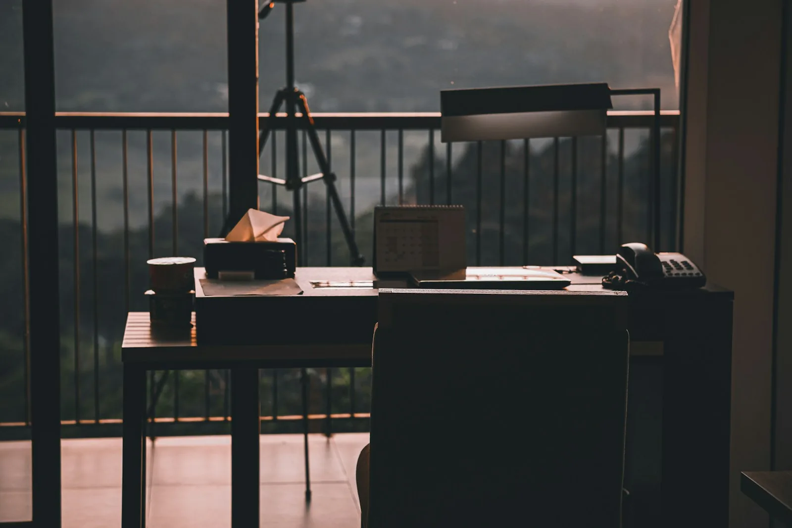 Empty desk at dawn by a balcony window, with a desk calendar, a desk phone and a lamp waiting for the day to start