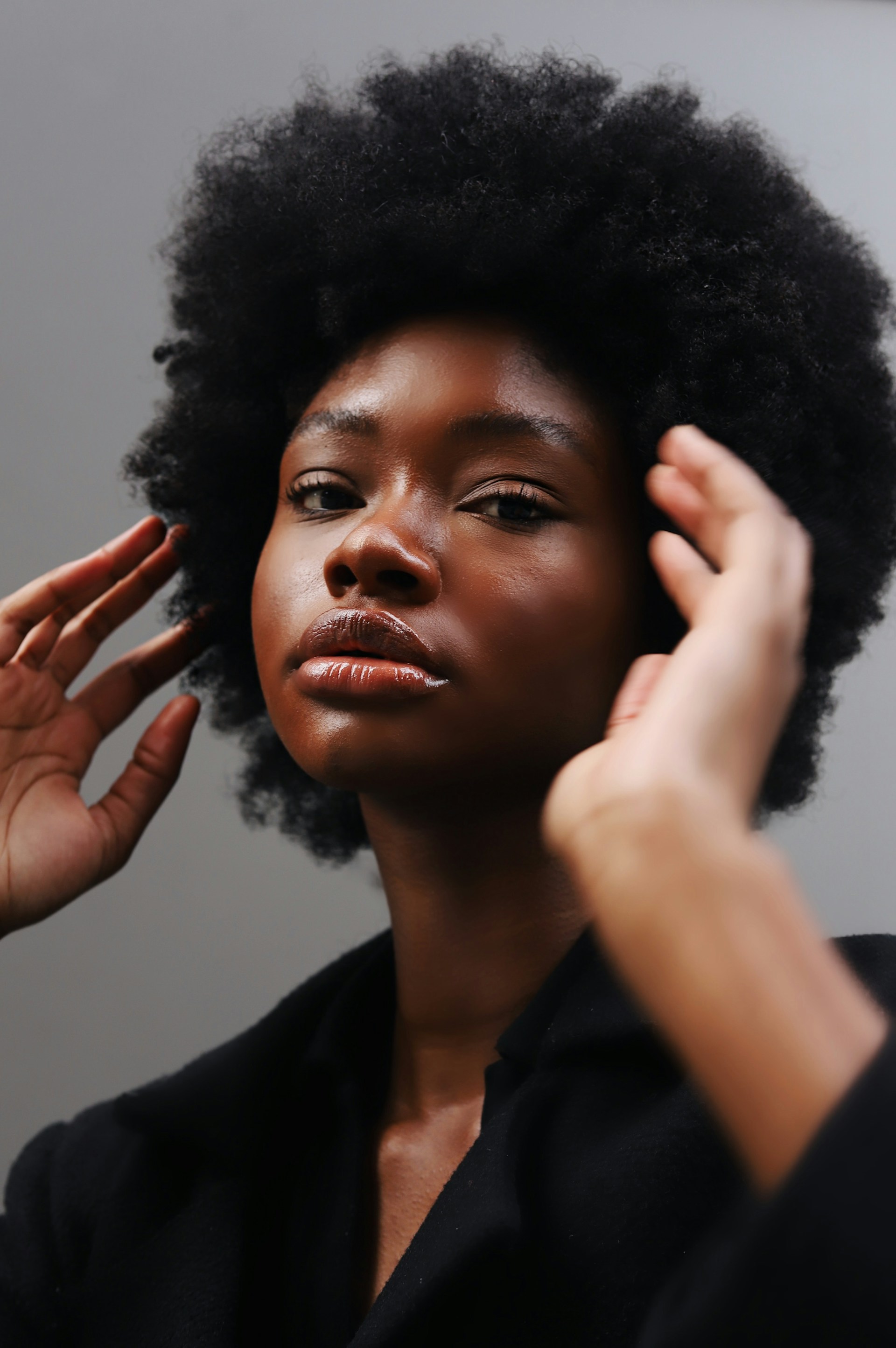 A portrait of a Black woman with a voluminous afro, posing with her hands near her face and looking directly at the camera with a neutral expression.