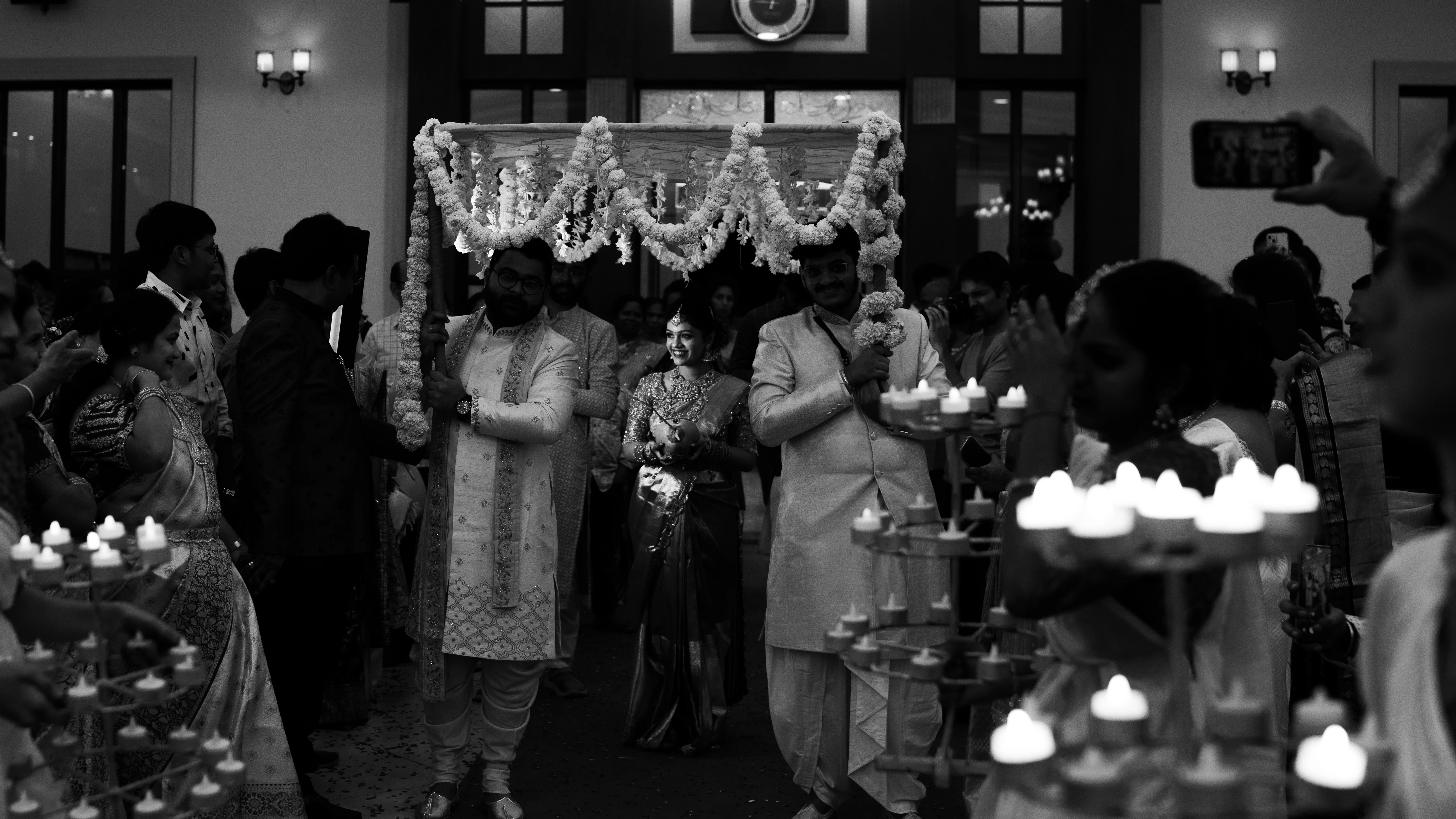 Fine art wedding photography in Hyderabad by Out of The Blues – black and white image of a bride walking down the aisle, surrounded by guests and flickering candles, creating a cozy atmosphere.