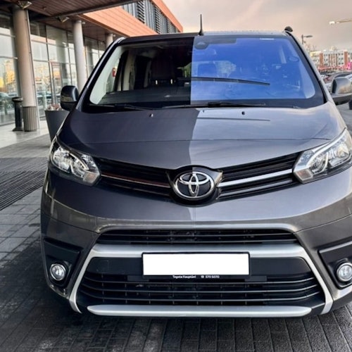Vista frontal de una furgoneta Toyota gris estacionada en un entorno urbano al aire libre, con edificios y una acera en el fondo.