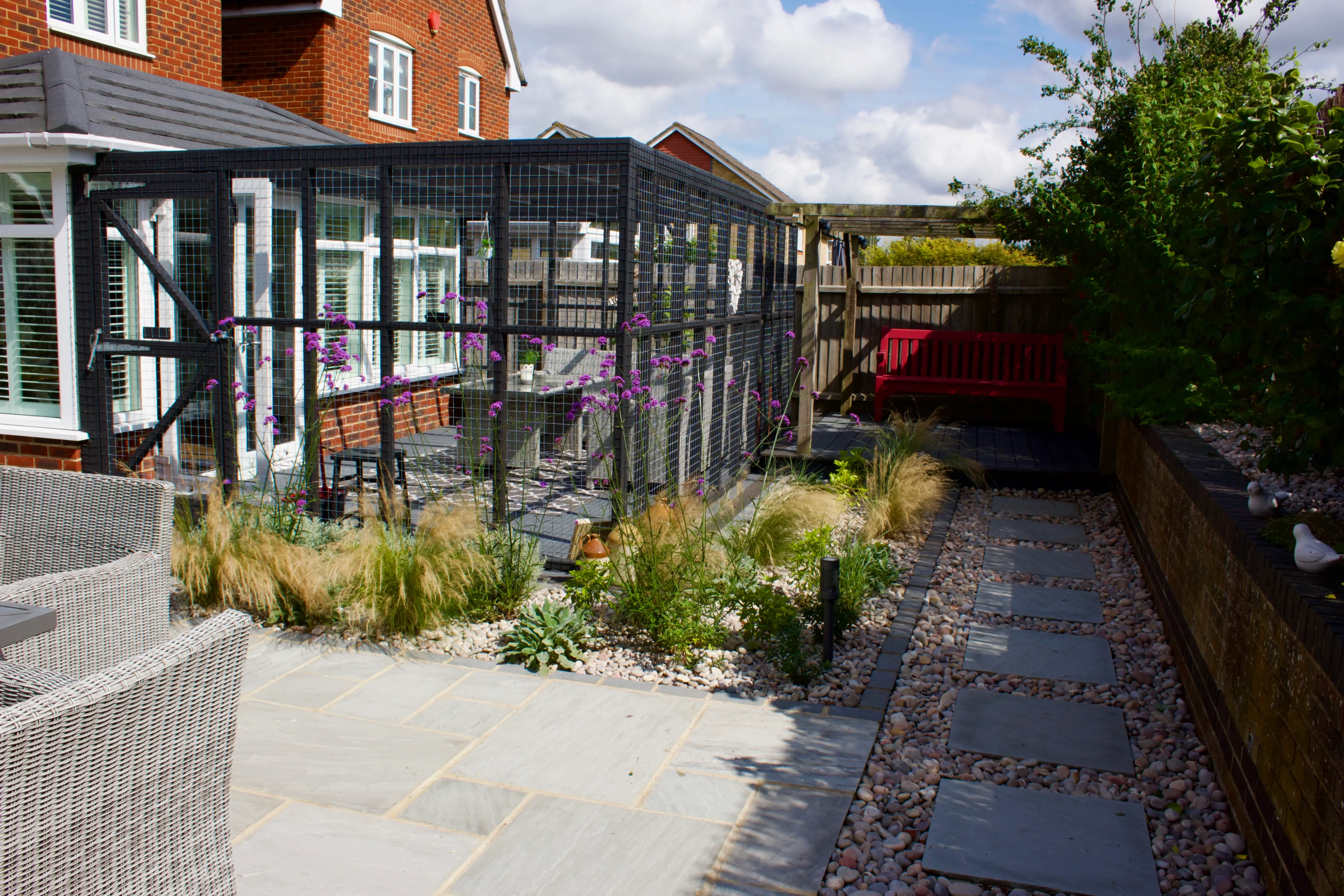 A landscaped garden with a pathway, flowers, and a glass structure in the background on a sunny day.