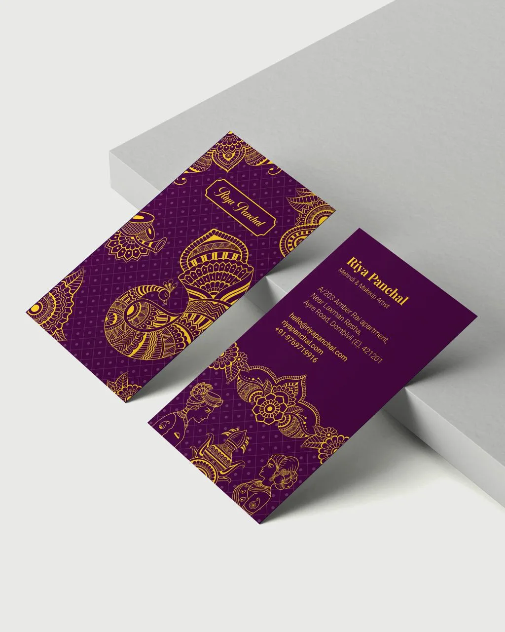 Two purple Indian wedding invitation cards with ornate gold floral and paisley patterns, displayed on a minimalist gray surface.